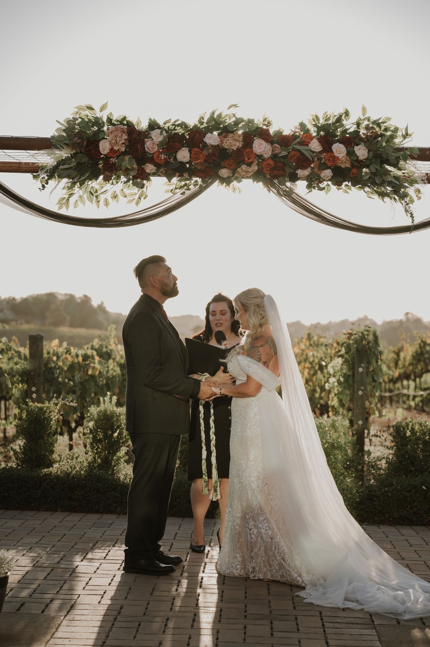Wedding ceremony under a floral arch in a vineyard during golden hour.