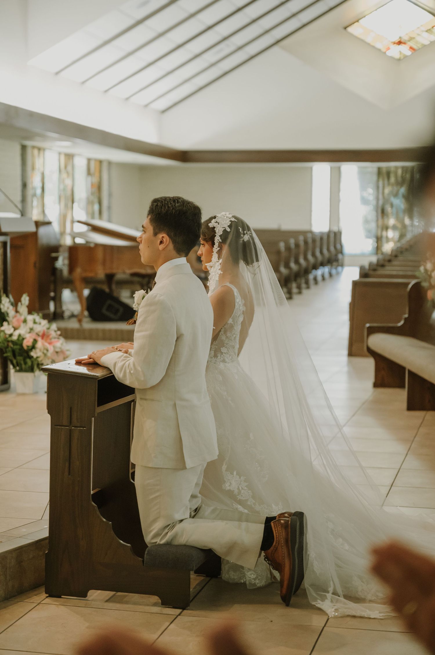 Bride and groom kneel together in prayer at wooden kneeler during intimate church wedding ceremony.