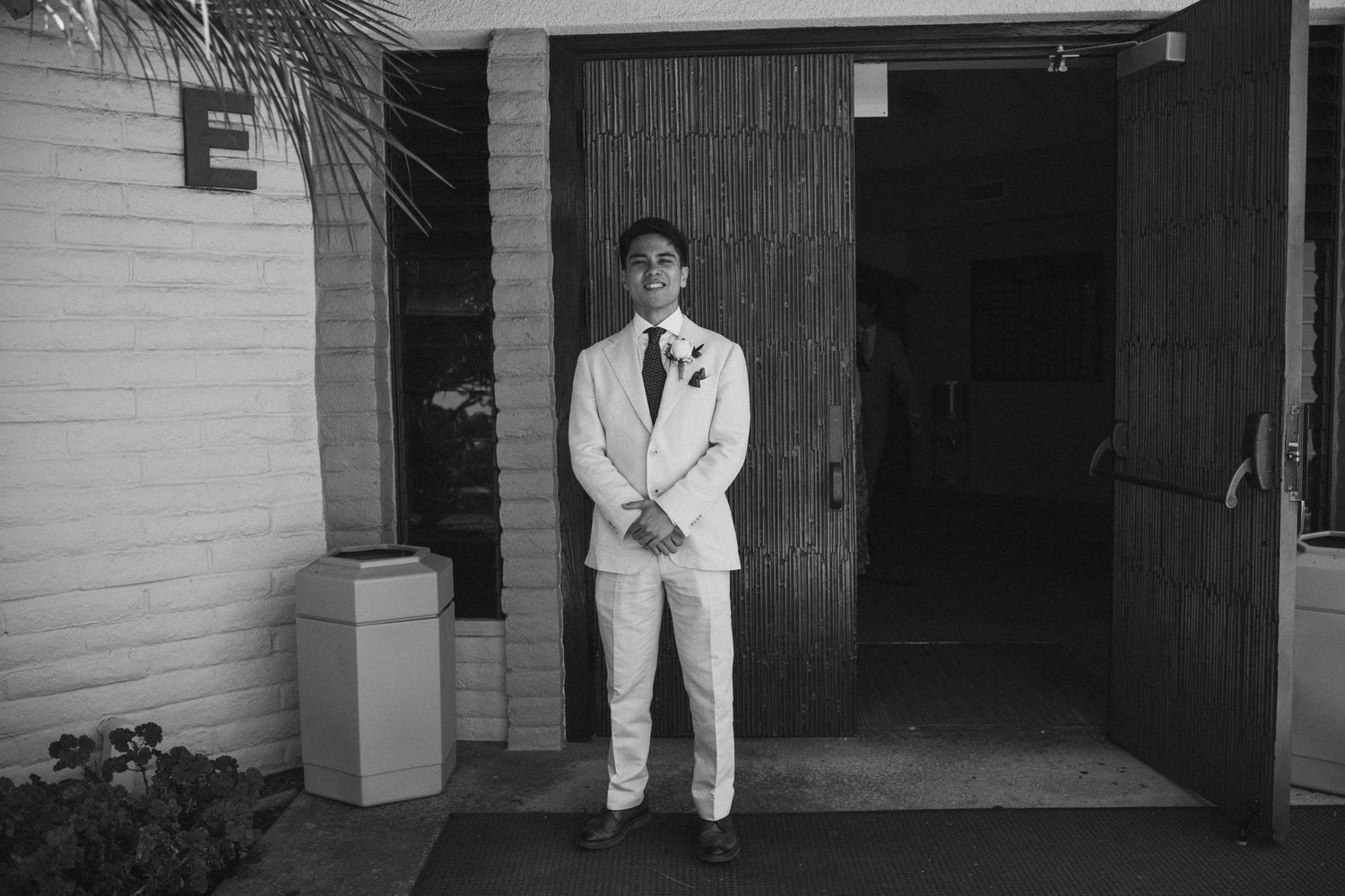 Groom in light suit with boutonniere stands confidently outside venue entrance in elegant black and white wedding photo.
