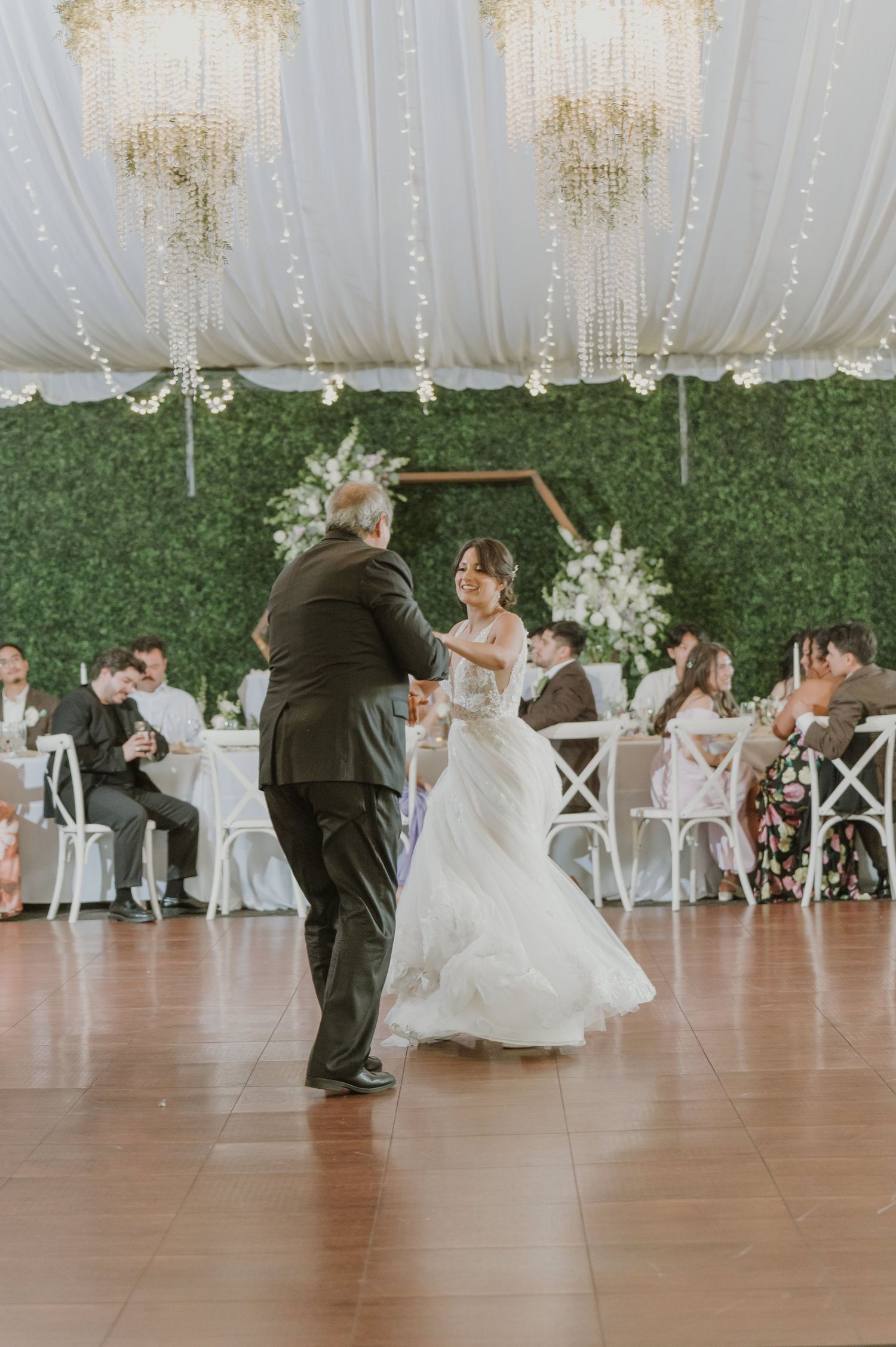 Bride in tulle gown shares father-daughter dance under gold chandeliers at elegant wedding reception.