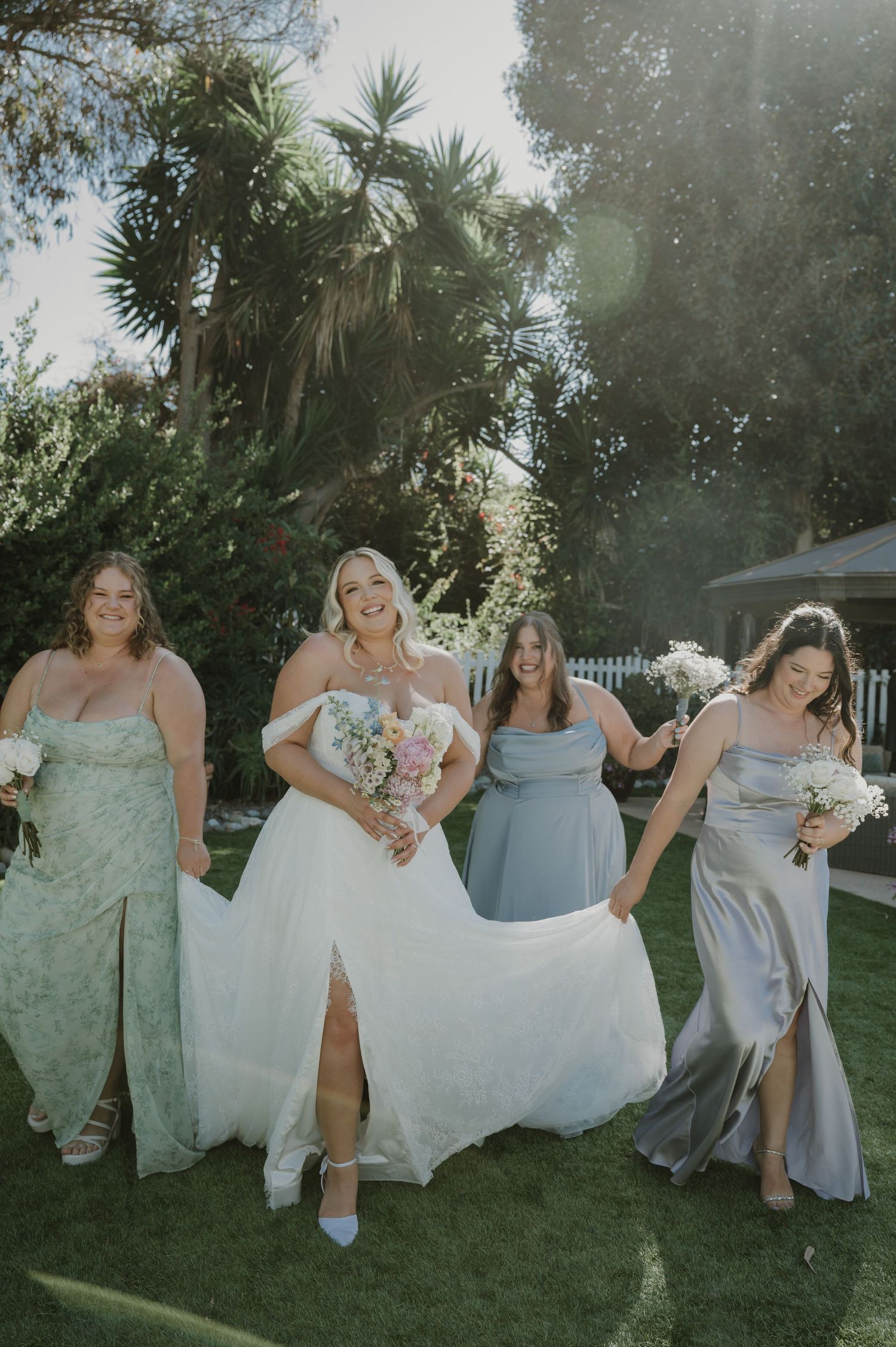 Bride in white gown with bridesmaids in sage and blue dresses walking outdoors in sunny garden setting.