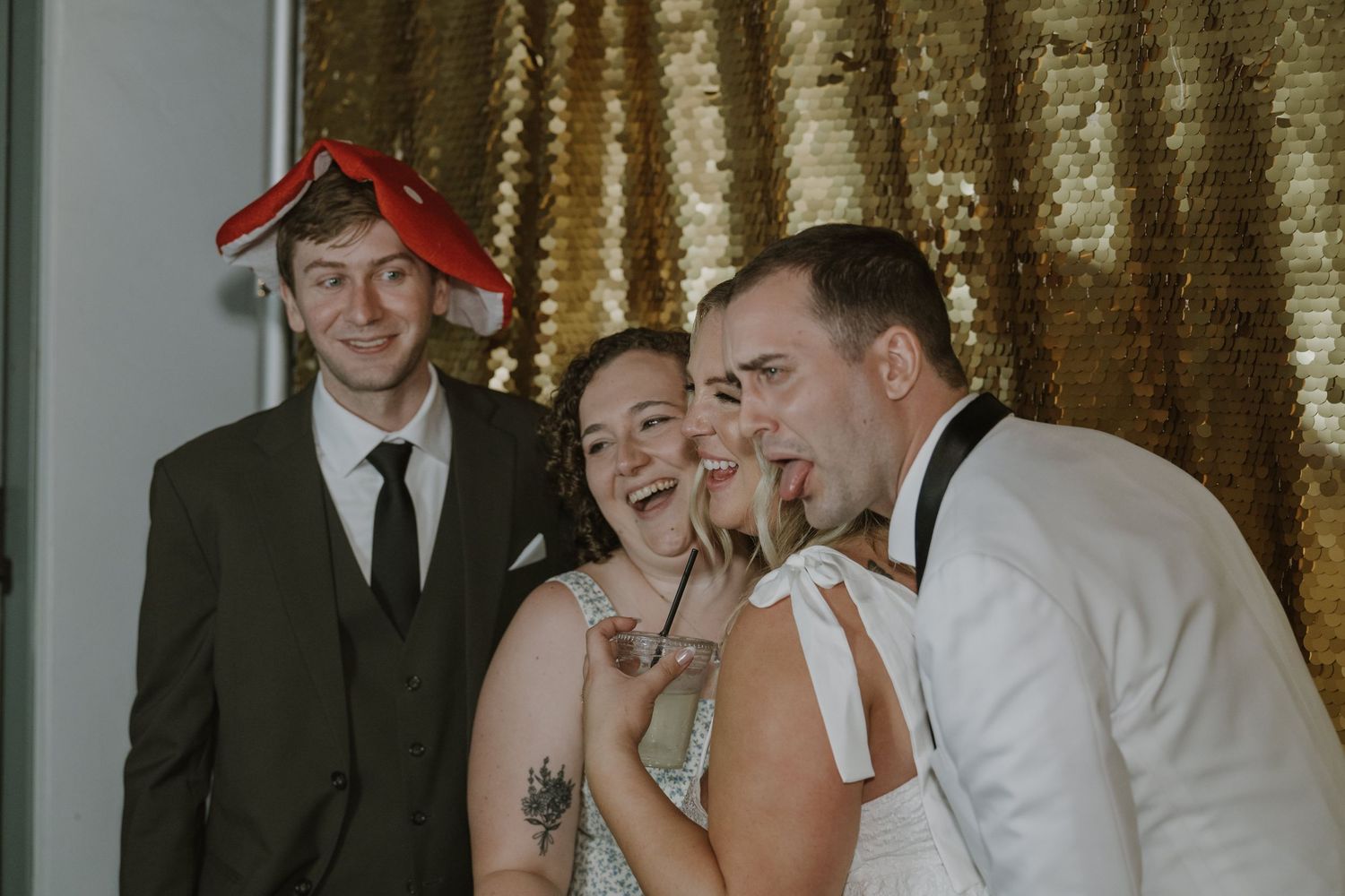 Bride, groom, and wedding guests laughing and posing together in front of a gold sequin photo booth backdrop.
