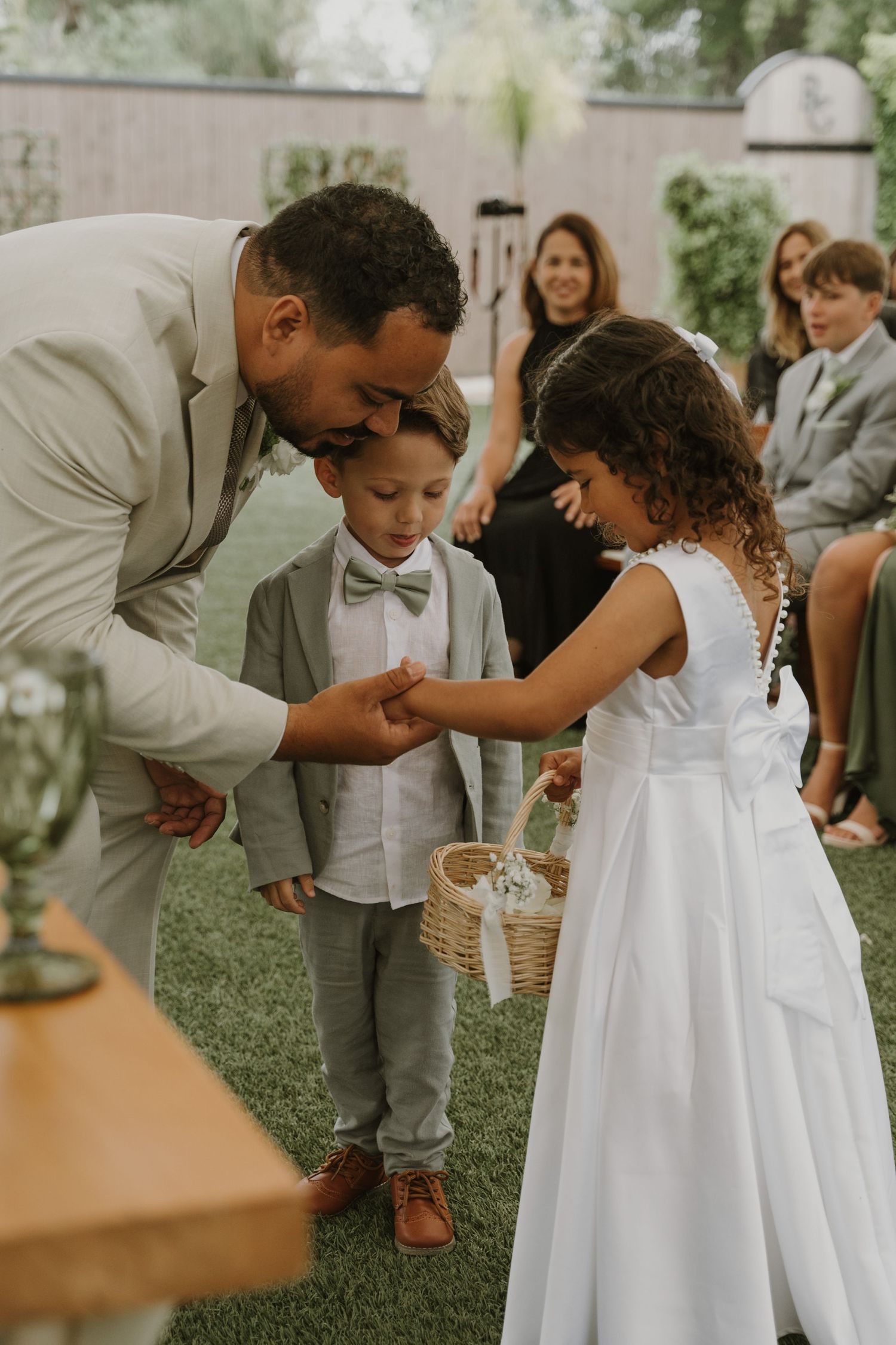 Groom in light gray suit greets ring bearer and flower girl with basket during outdoor wedding ceremony on green lawn.