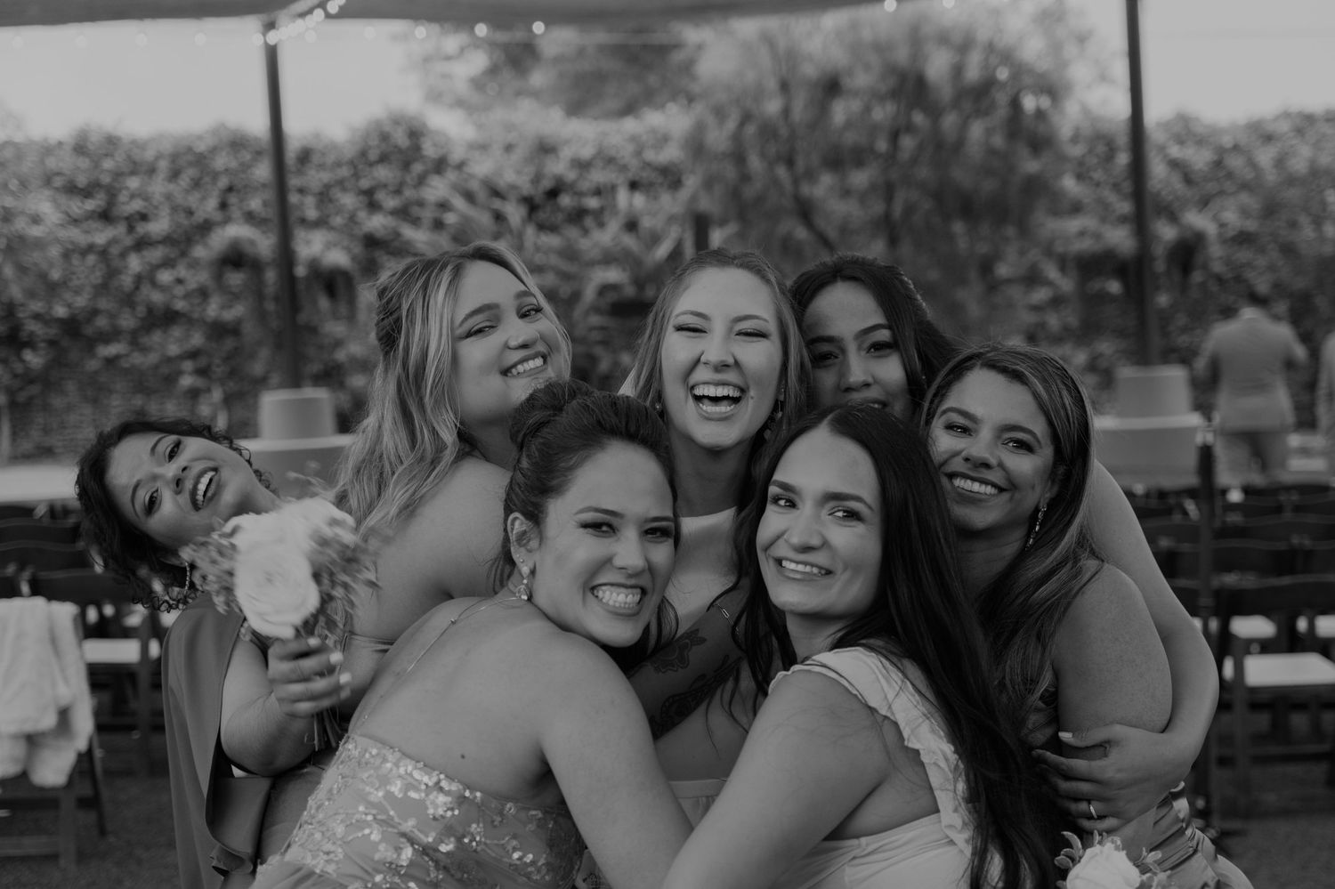 Six women in formal attire laughing and hugging together at an outdoor wedding celebration in black and white photo.