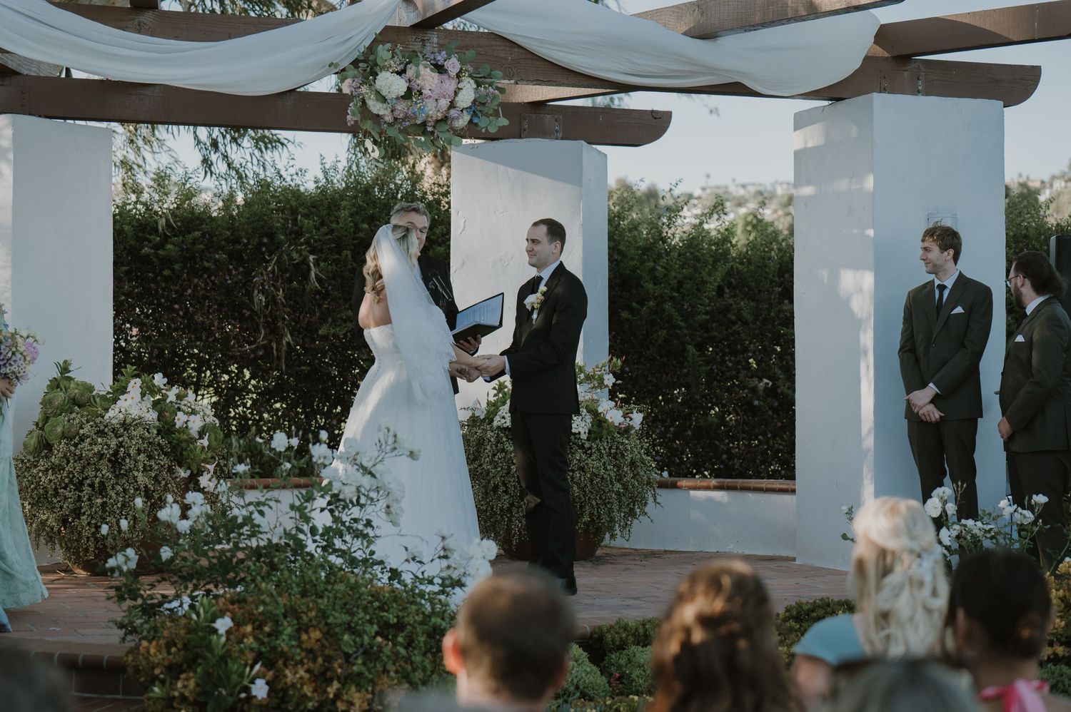 Outdoor wedding ceremony under a pergola with floral arch, bride and groom exchanging vows before seated guests.