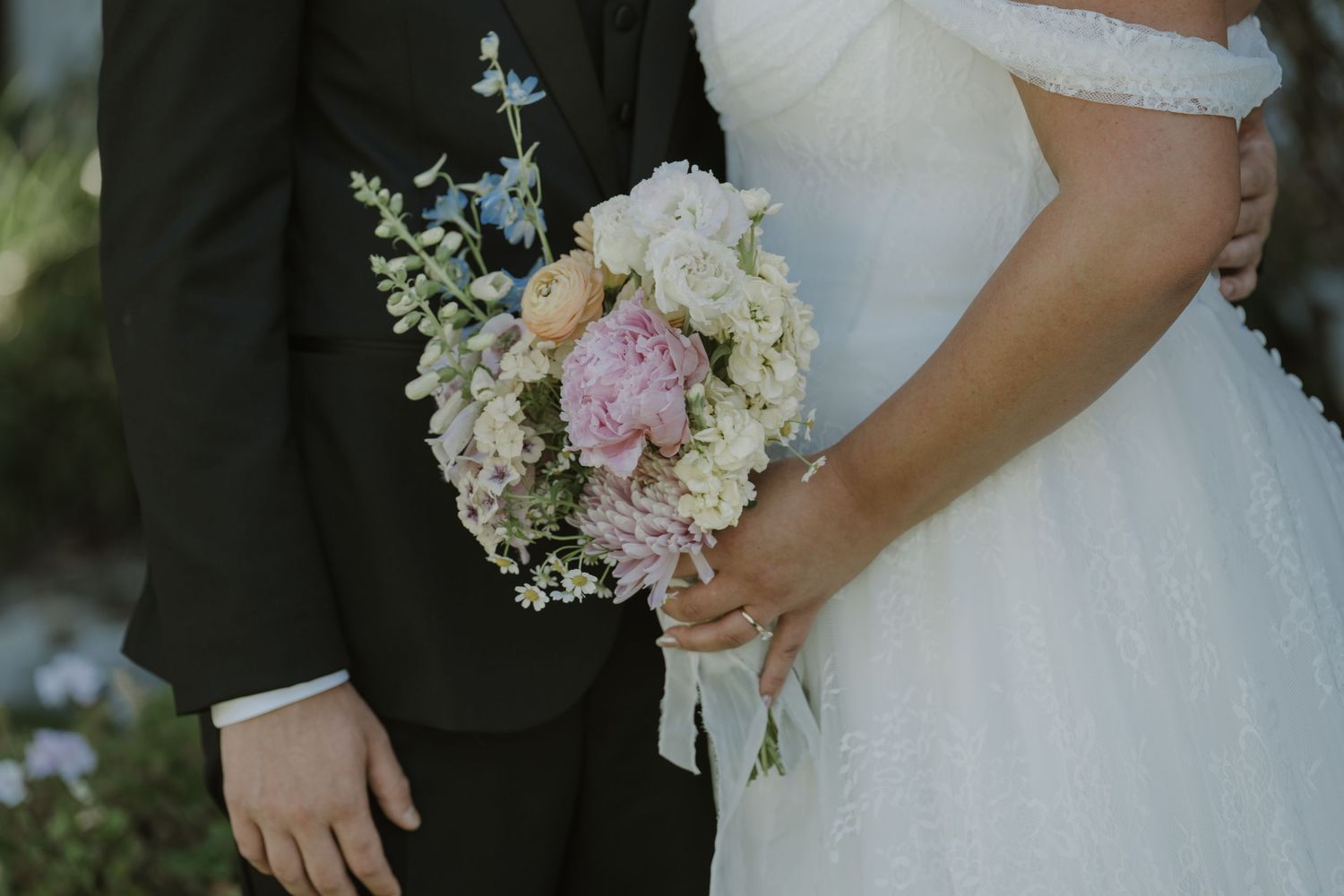 Bride and groom holding a pastel pink and white floral bouquet with greenery on their wedding day.