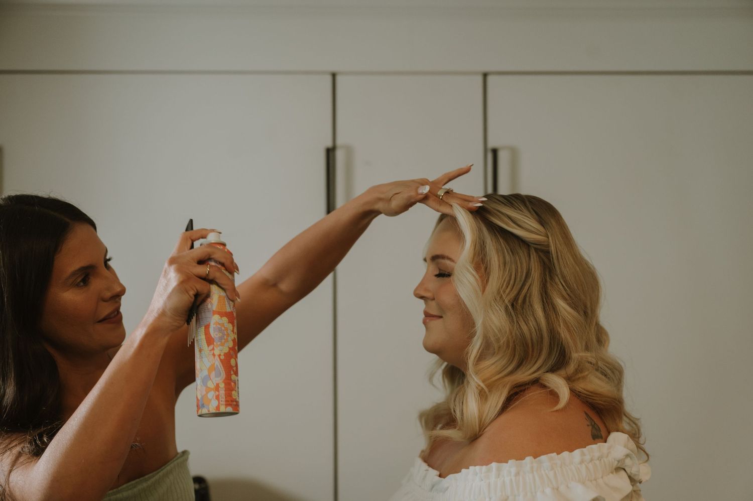 A hairstylist applying hairspray to a blonde bride with curly hair during bridal hair preparation.
