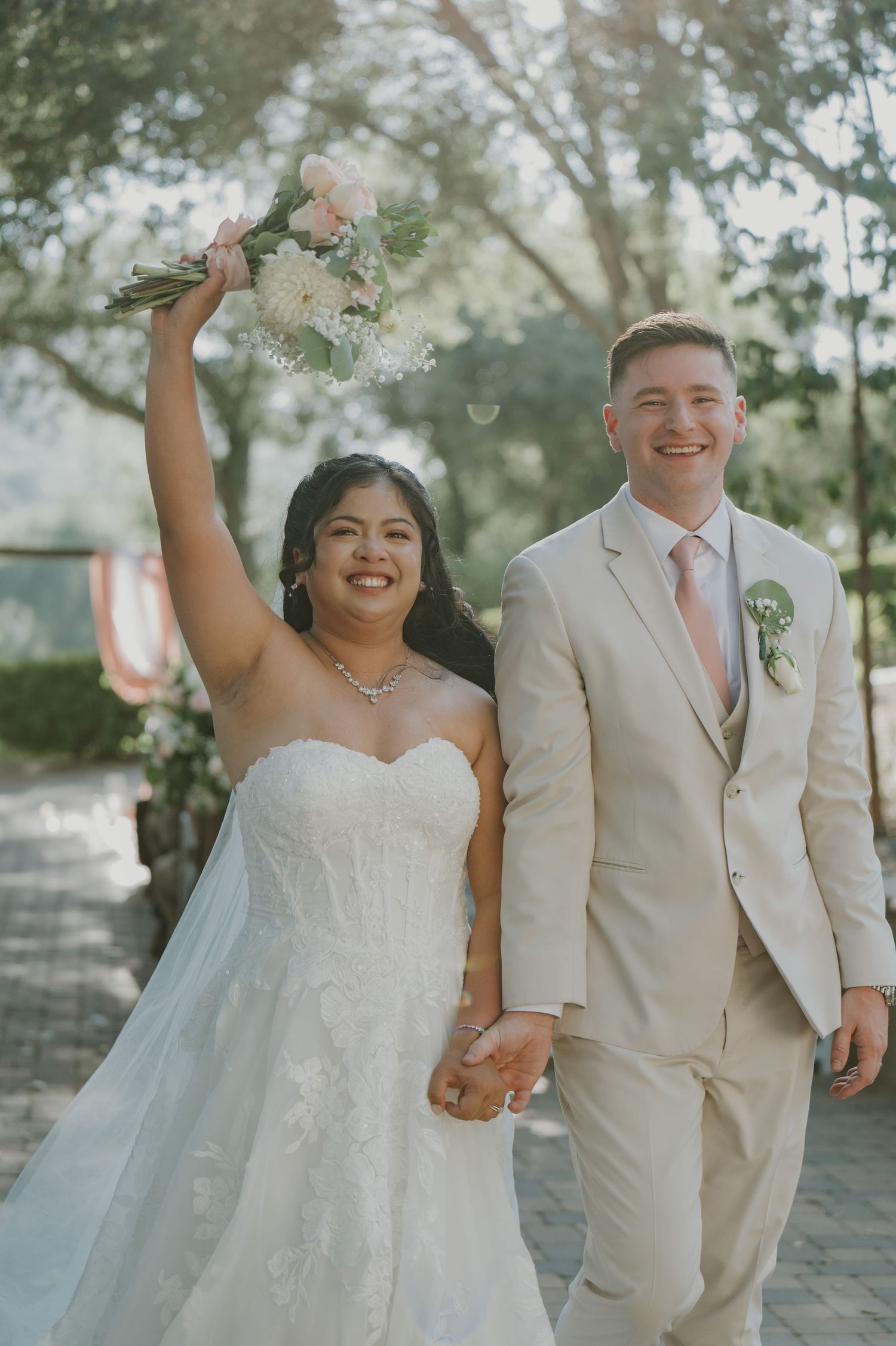 Joyful newlyweds celebrate after ceremony, bride raises bouquet as groom smiles in beige suit.
