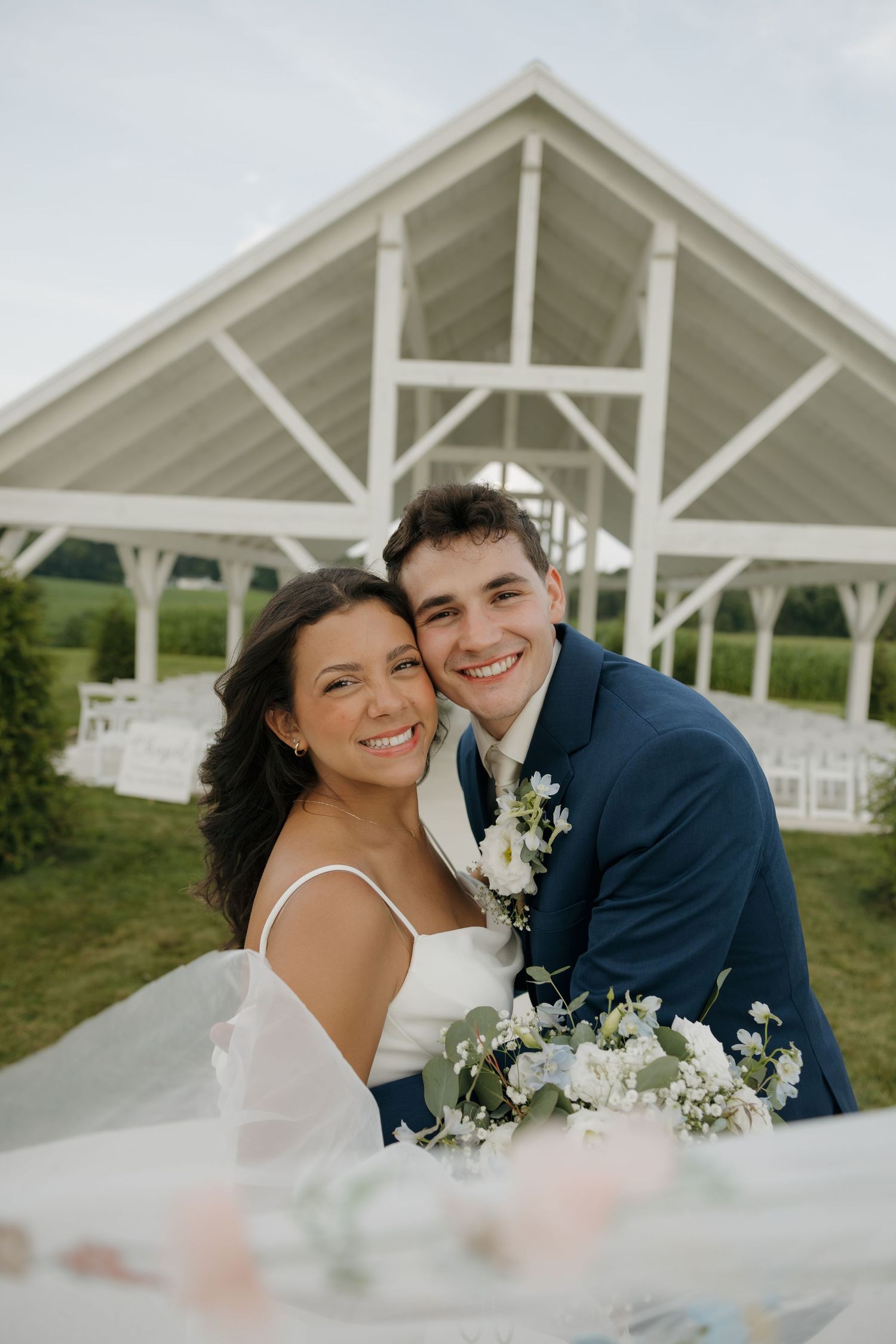 Smiling bride and groom pose together in front of a white outdoor wedding chapel.