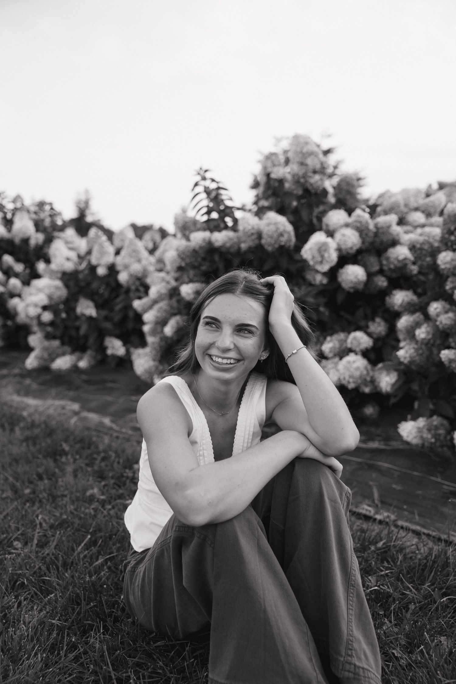 Black and white portrait of a smiling woman sitting on grass in front of blooming hydrangea bushes.