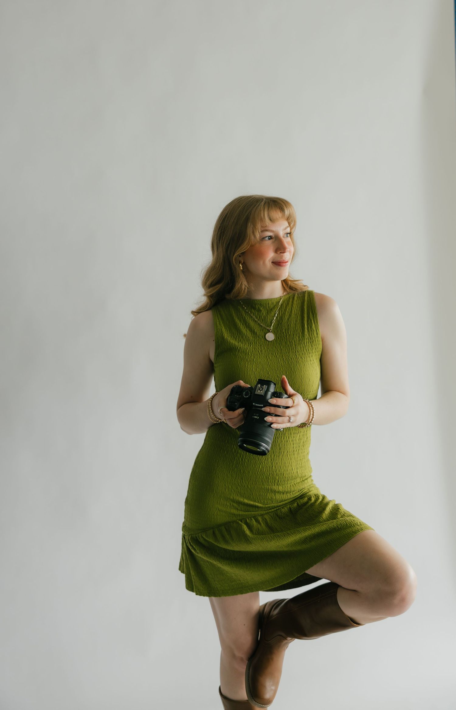 A young woman in a green sleeveless dress holds a camera while posing gracefully on one leg against a white background.