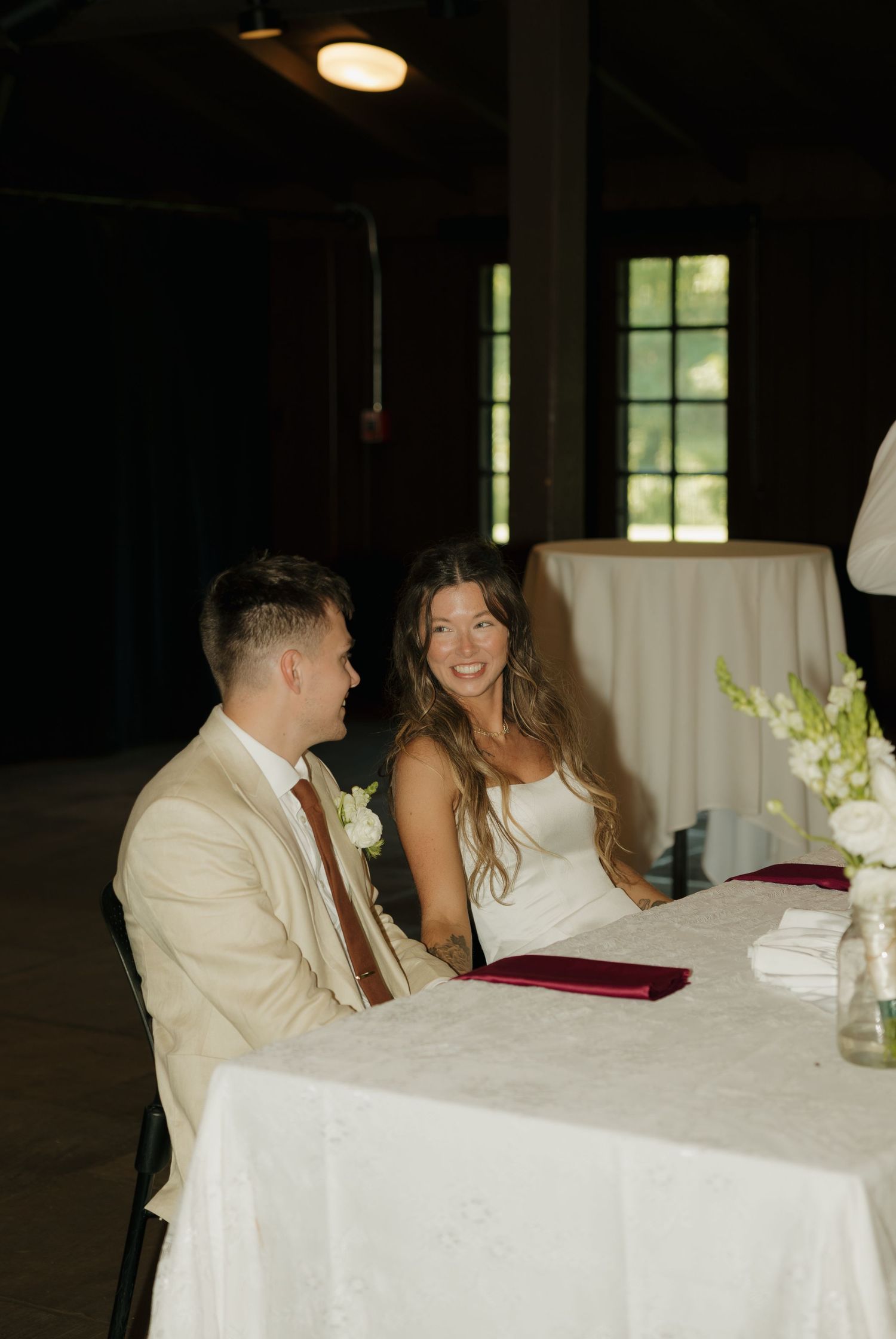 Bride and groom smiling at sweetheart table during wedding reception in rustic venue with white floral arrangements.