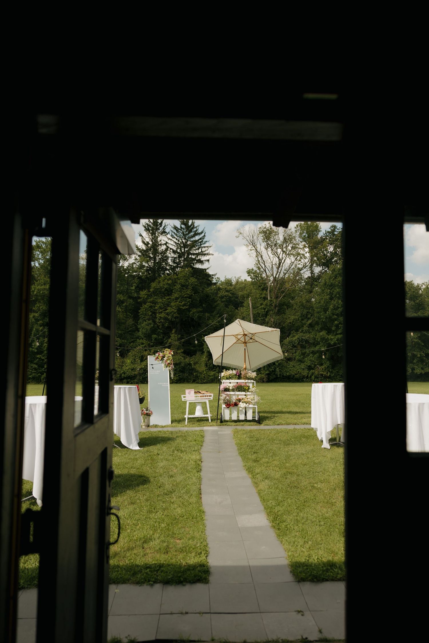 Elegant outdoor wedding ceremony setup viewed through open doors, featuring white draped tables, a stone pathway, and patio umbrella.