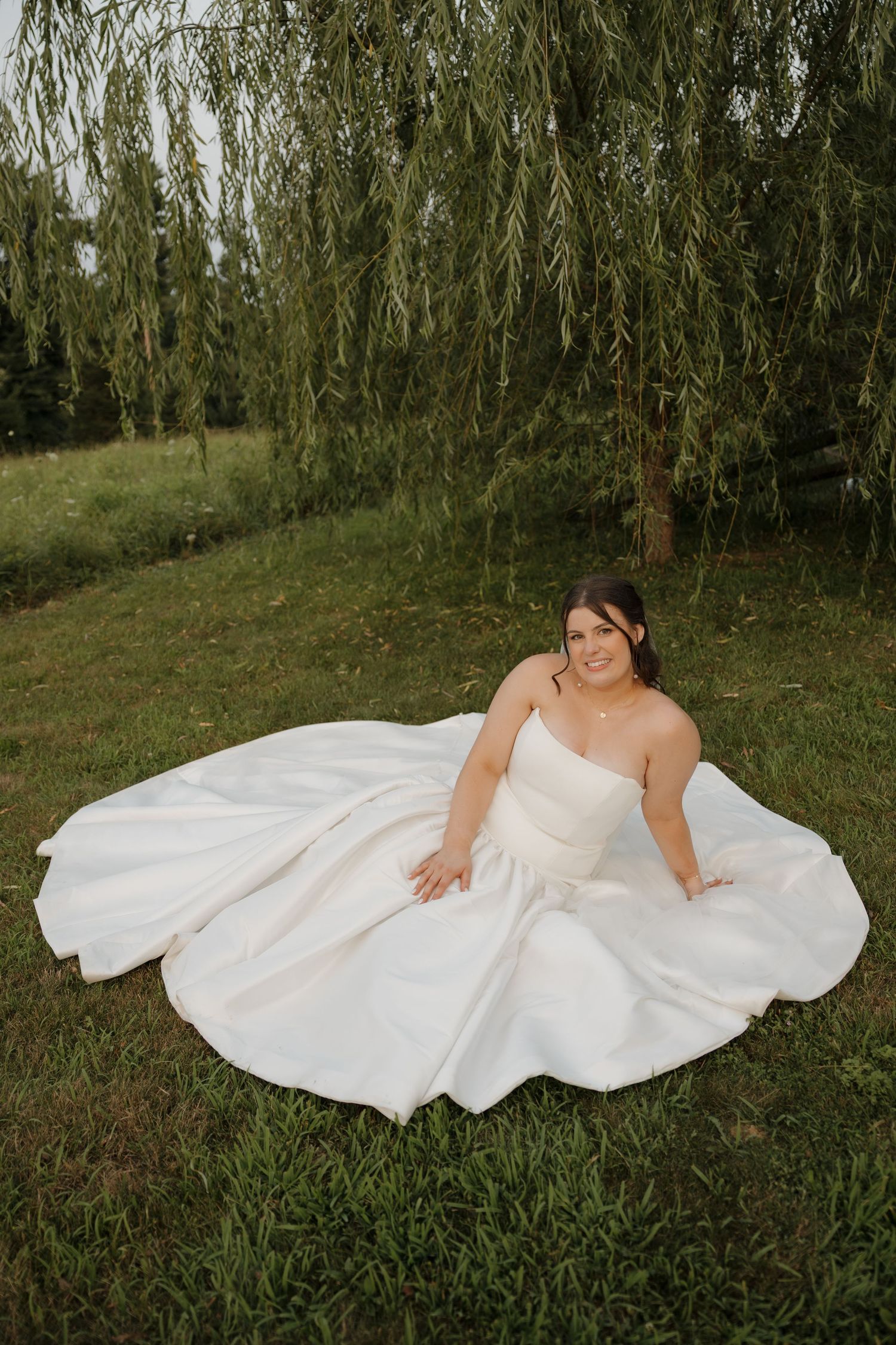 Bride in a flowing white ballgown sitting on lush grass beneath a weeping willow tree, smiling softly.