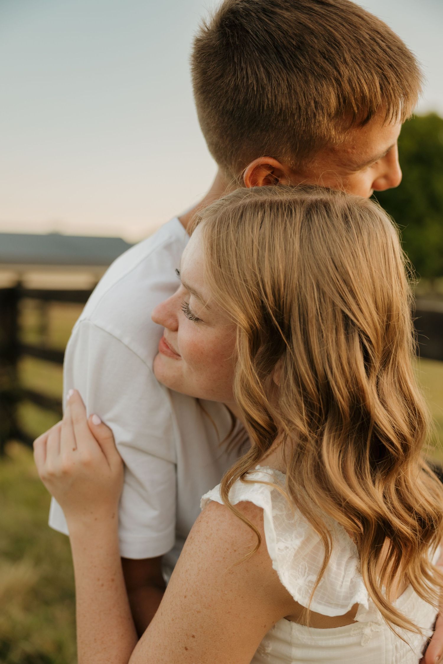A young couple embraces outdoors at golden hour, the woman smiling as she leans against the man in a white shirt.