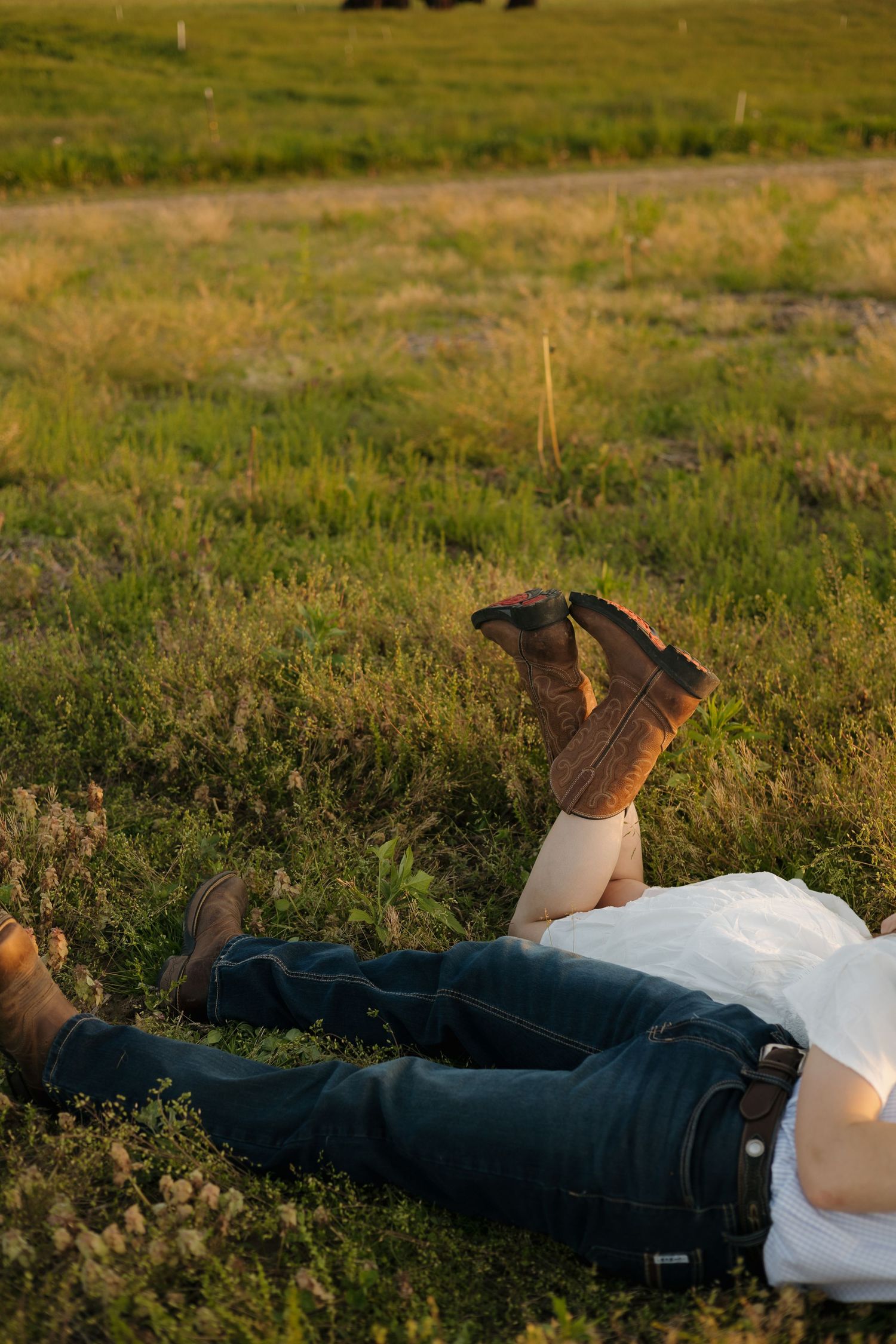 Couple lying in grass field at sunset, woman in white dress and cowboy boots, man in jeans and white shirt.