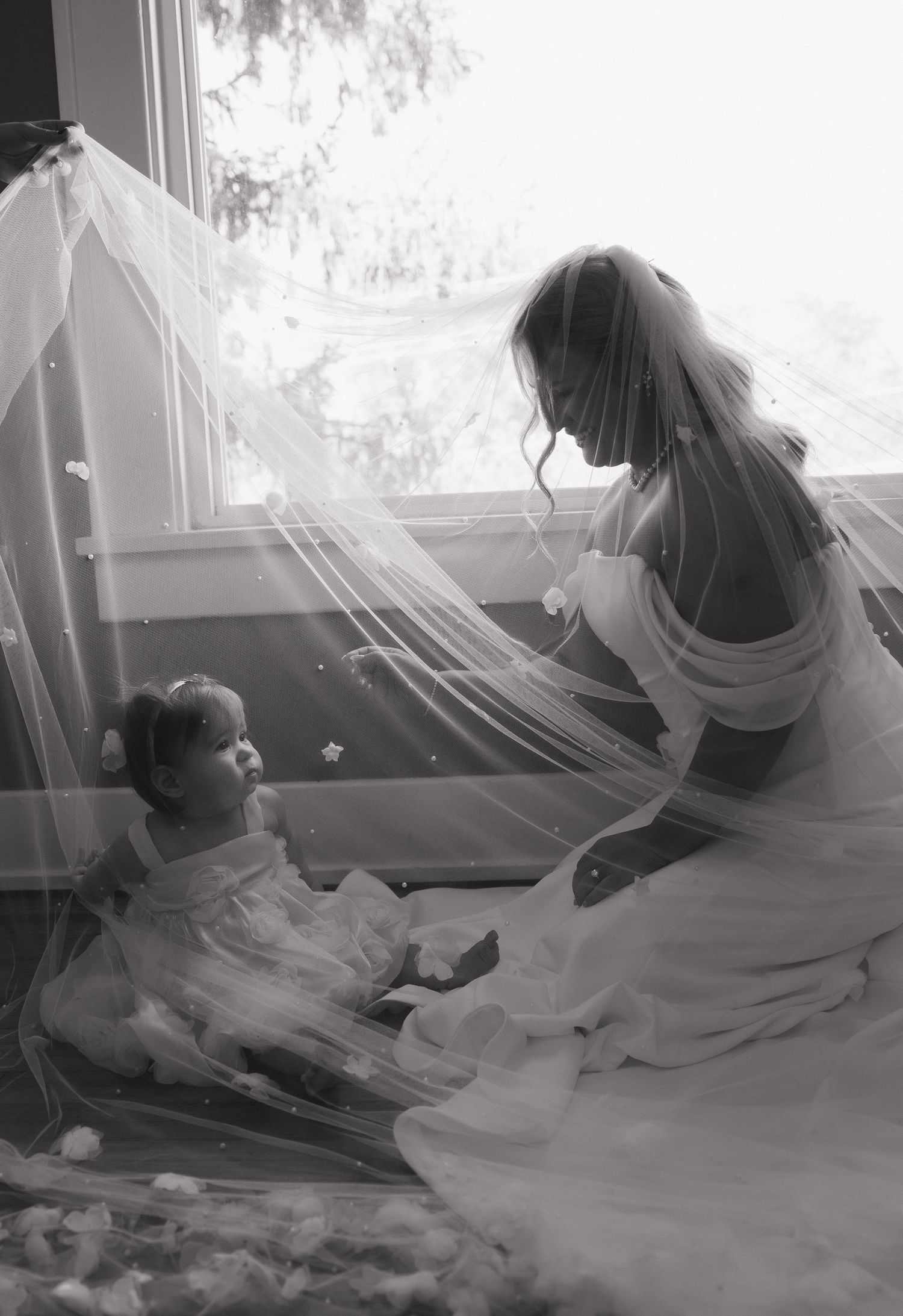 Black and white photo of a bride in a veil kneeling beside a young flower girl by a sunlit window.