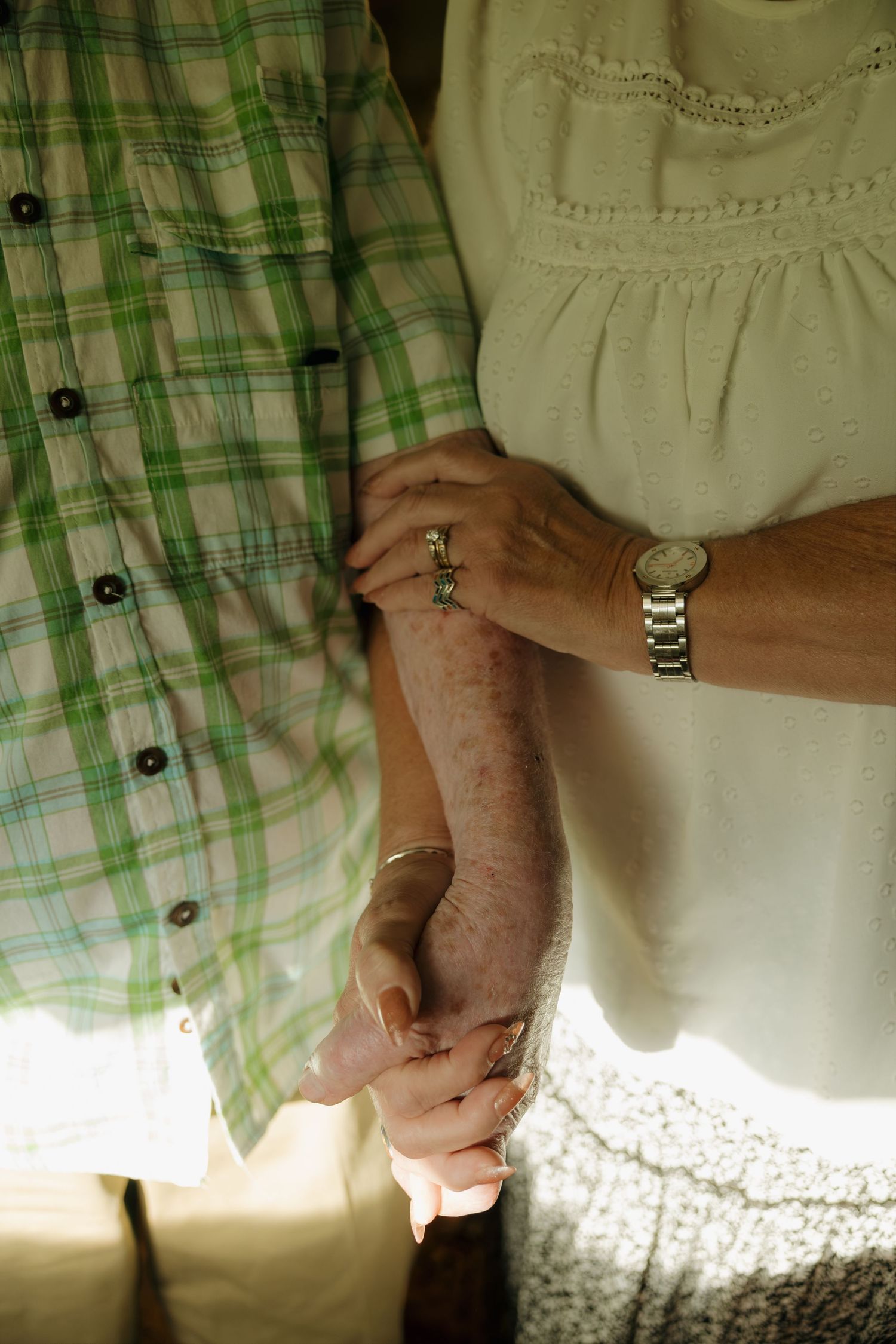 Close-up of elderly couple holding hands, woman wearing a watch and ring, man in plaid shirt.