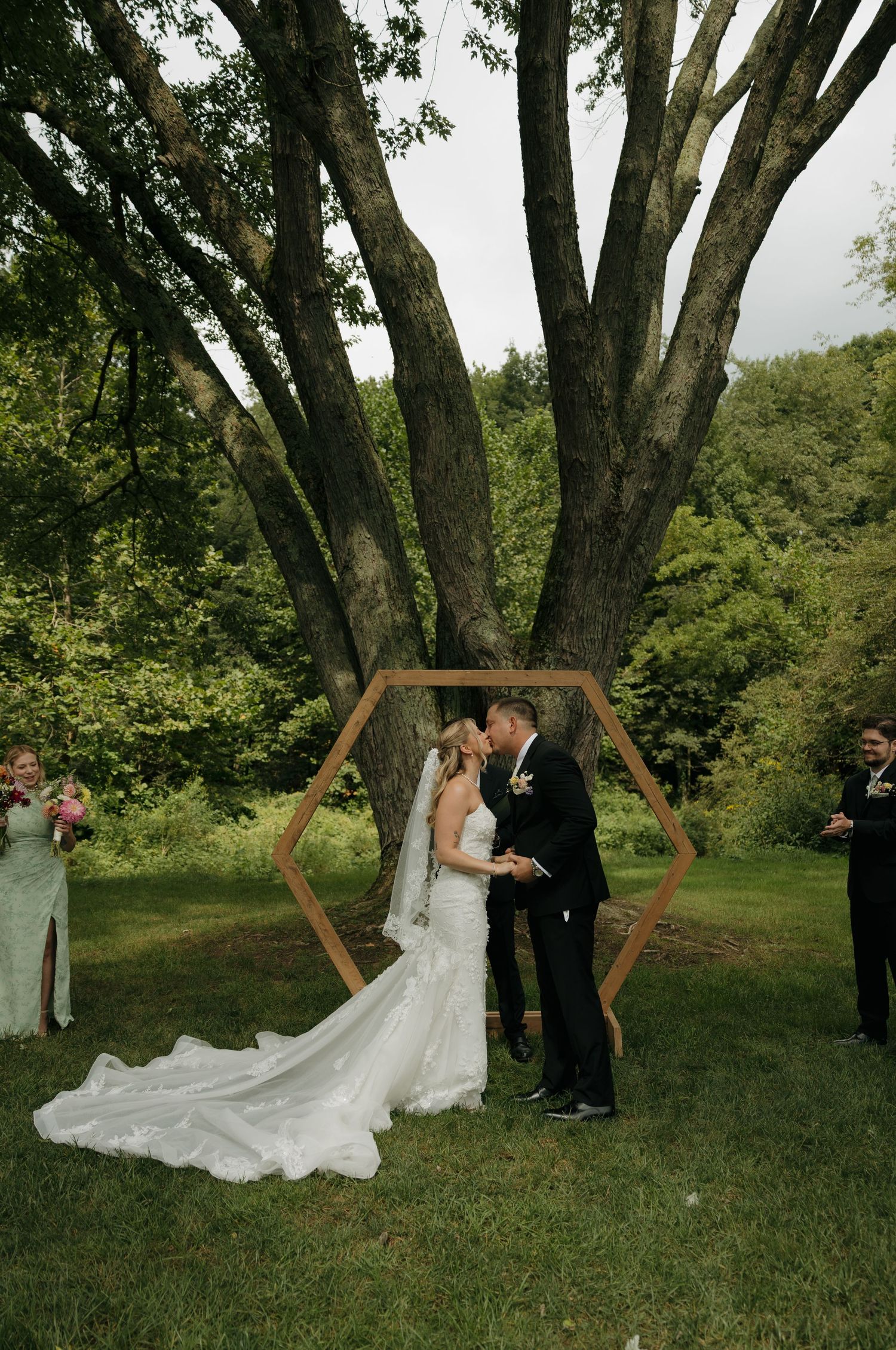 Bride and groom share first kiss under hexagonal wooden arch surrounded by lush green trees at outdoor wedding ceremony.