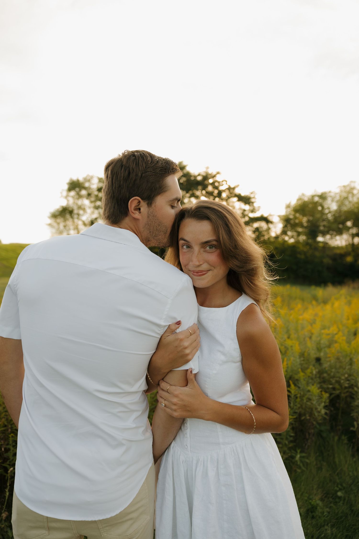Couple embracing in a golden field at sunset, woman in white dress smiling softly at camera.