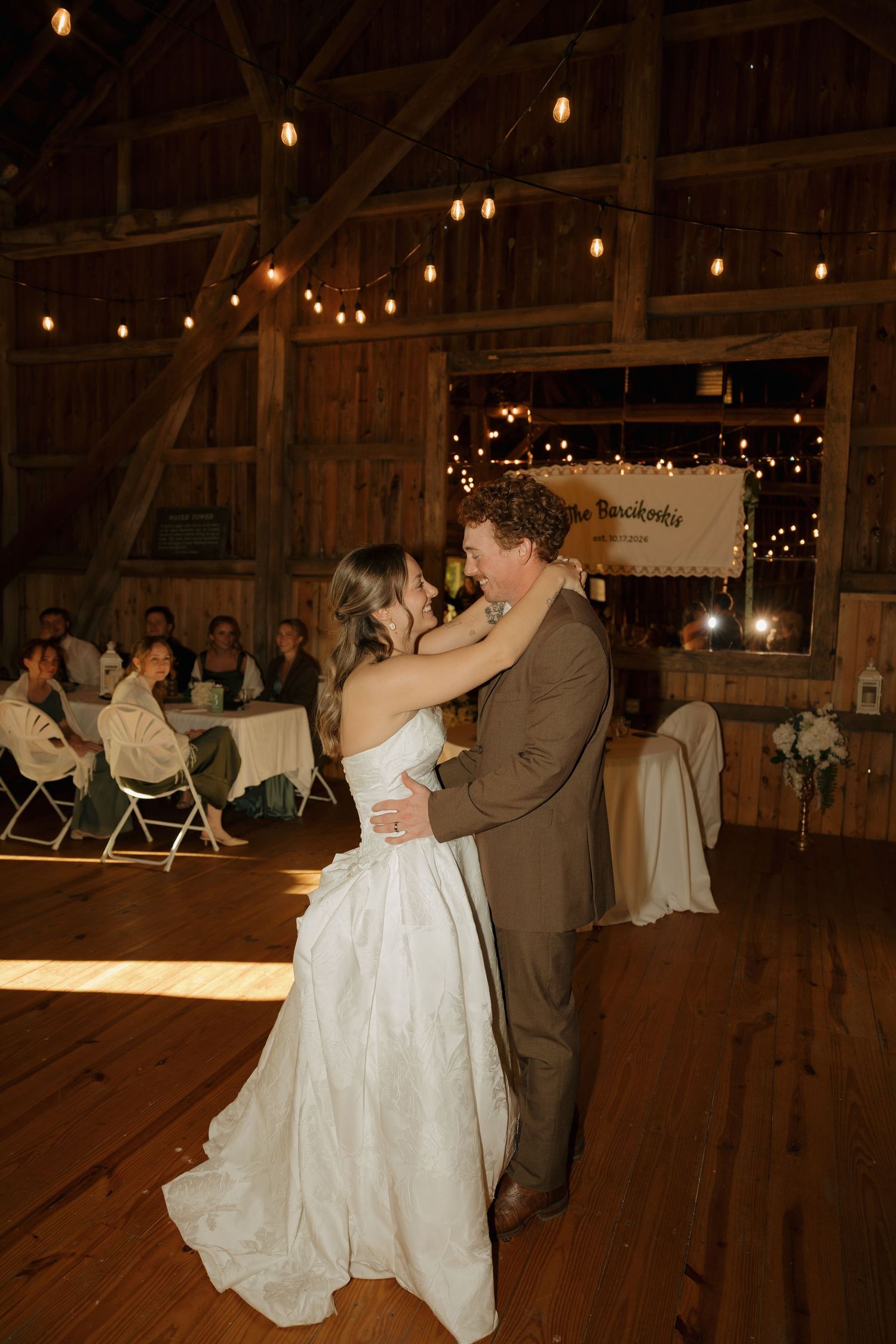 Bride in a white gown and groom in a brown suit share a first dance in a rustic barn wedding venue with string lights.