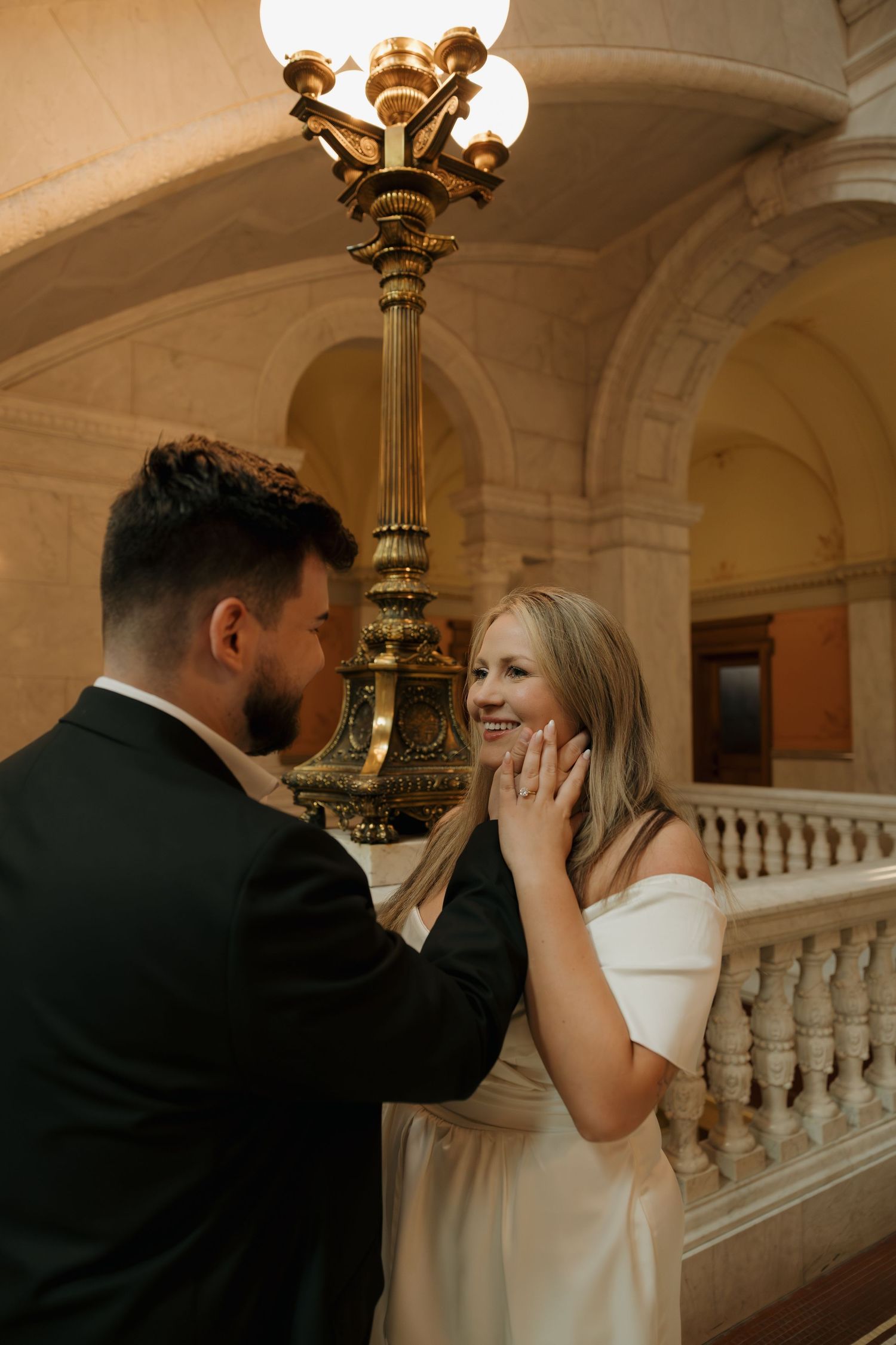 Couple sharing a tender moment in an elegant marble building, bride in white off-shoulder dress smiling as groom holds her face.