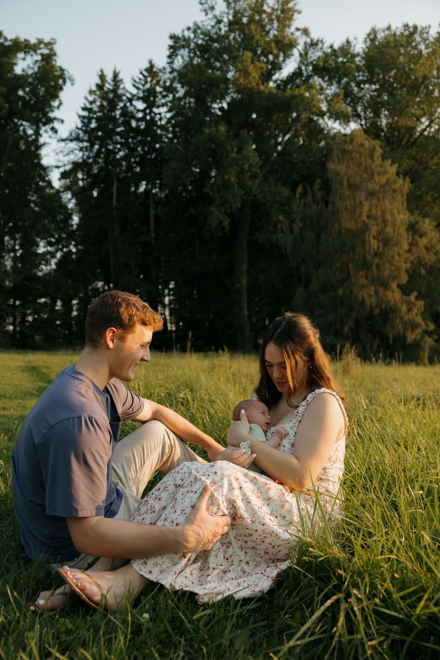 Young couple sitting in green meadow at golden hour, tenderly holding their newborn baby outdoors.