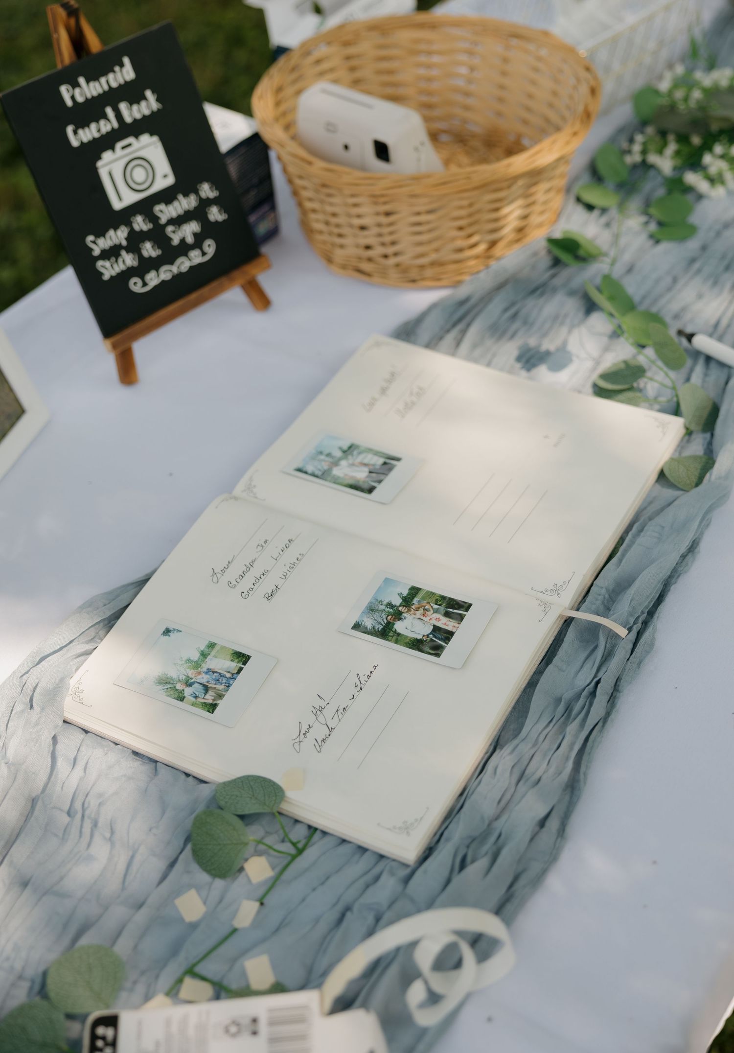 Polaroid guest book display with instant photos, wicker basket, and Instax camera on decorated wedding table.
