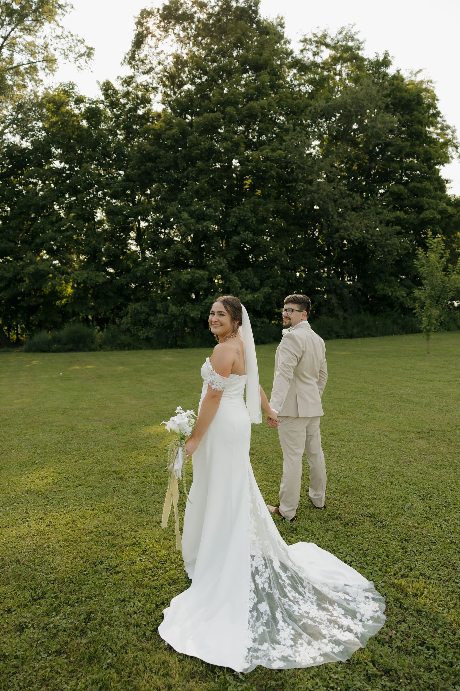 Bride in white off-shoulder gown with lace train holds bouquet, groom in tan suit, green trees backdrop.