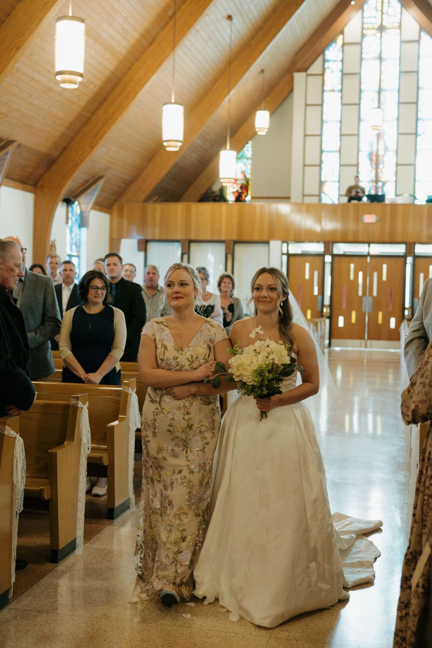 A bride in a white gown holding a bouquet walks arm in arm with a woman in a floral dress down the church aisle as guests watch.