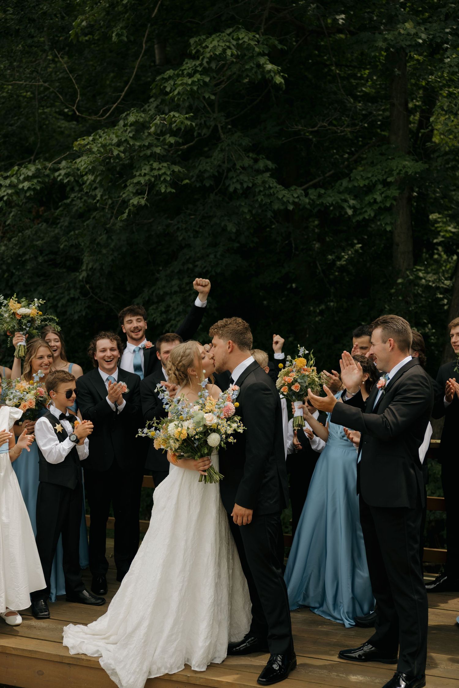 Bride and groom share a kiss surrounded by cheering wedding party in blue attire, holding colorful bouquets outdoors.