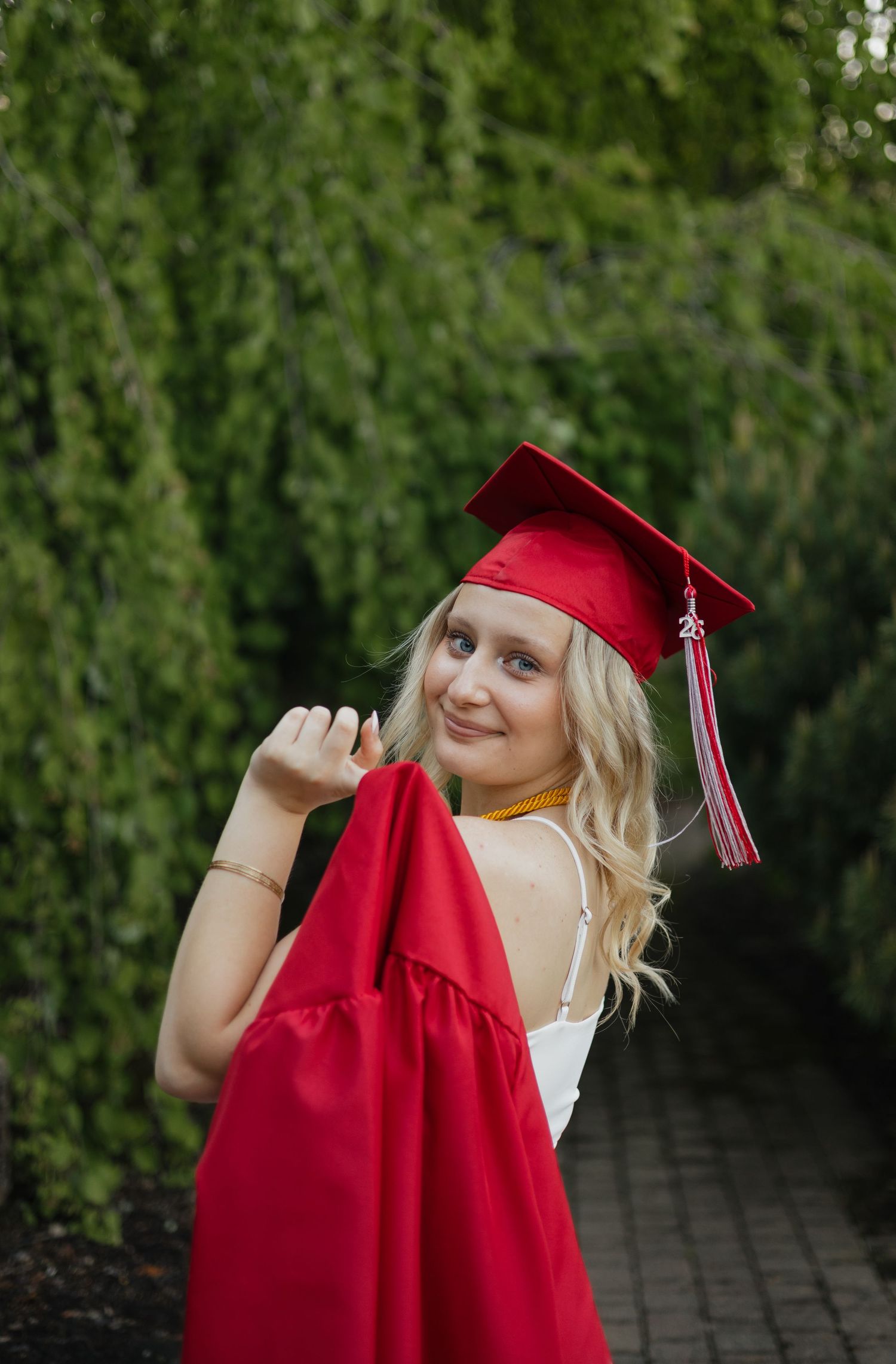 Smiling blonde graduate in red cap and gown poses on a tree-lined garden path.