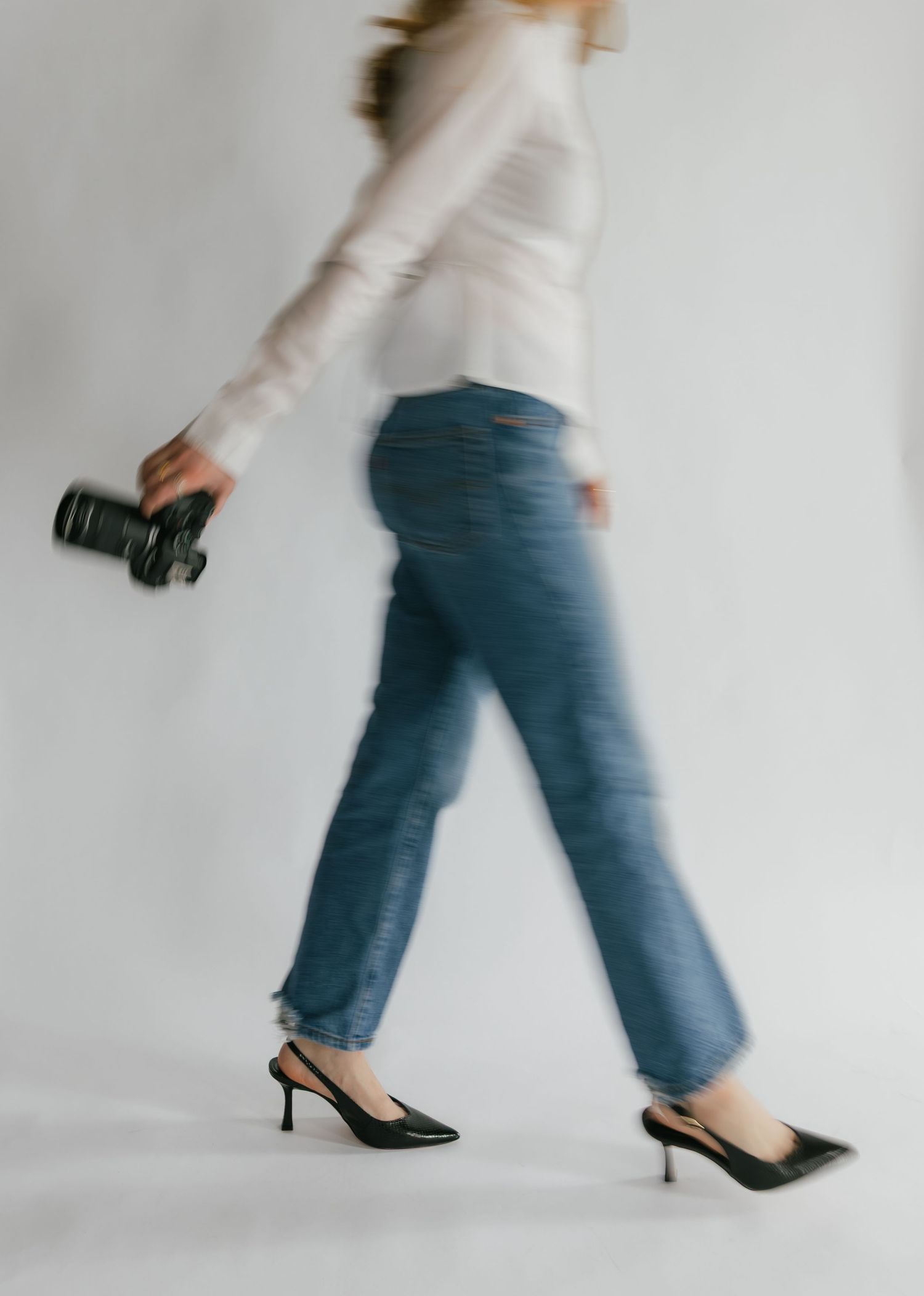 A woman in blue jeans and heels walks briskly while holding a camera, captured in a motion blur effect.