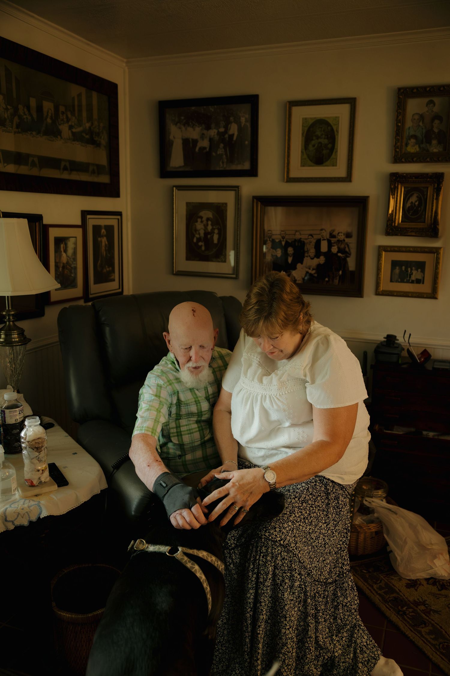 Elderly couple sitting together in armchair, examining hand, surrounded by family photos on wall in cozy living room.