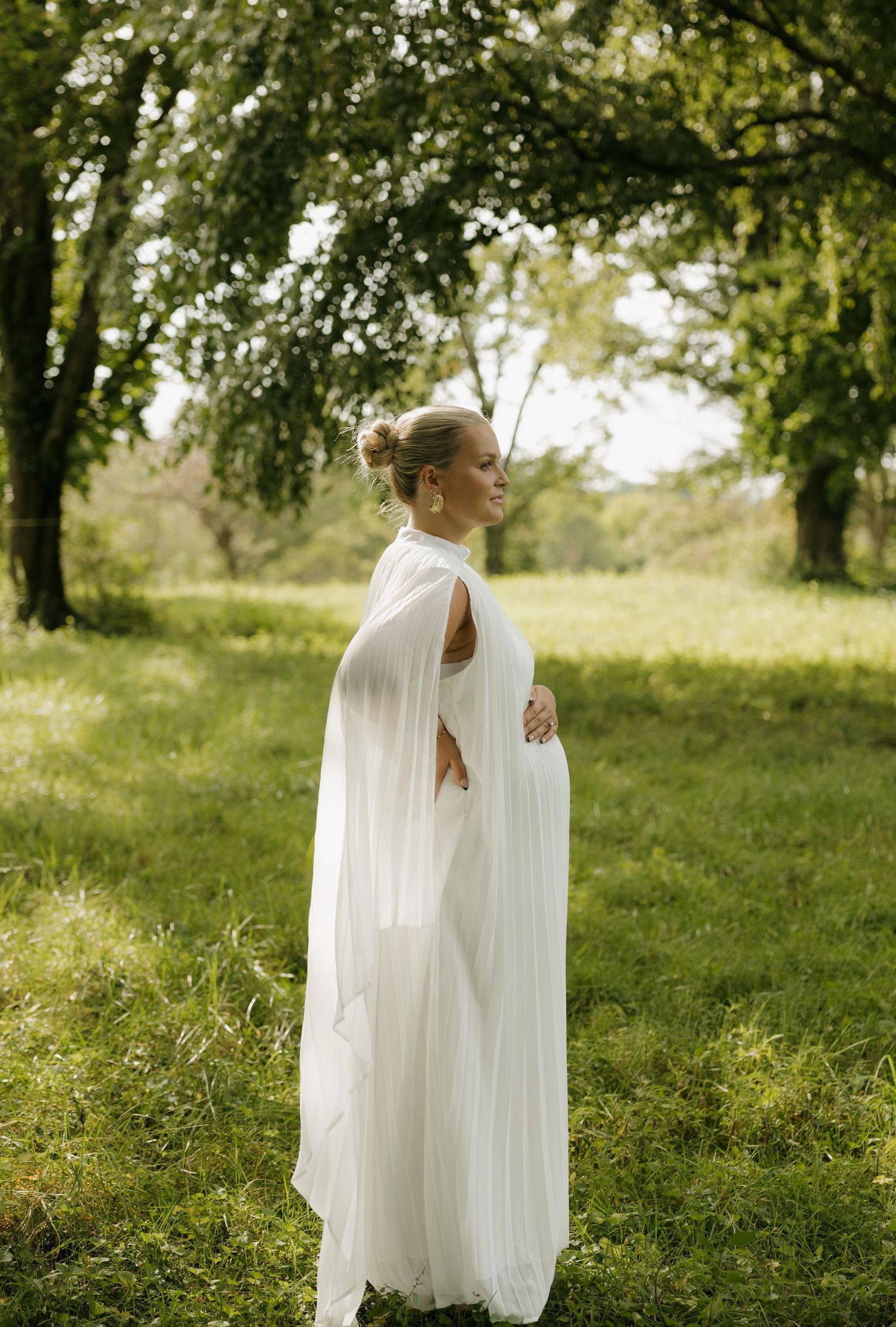 Bride in white flowing cape gown standing in lush green meadow surrounded by trees on her wedding day.