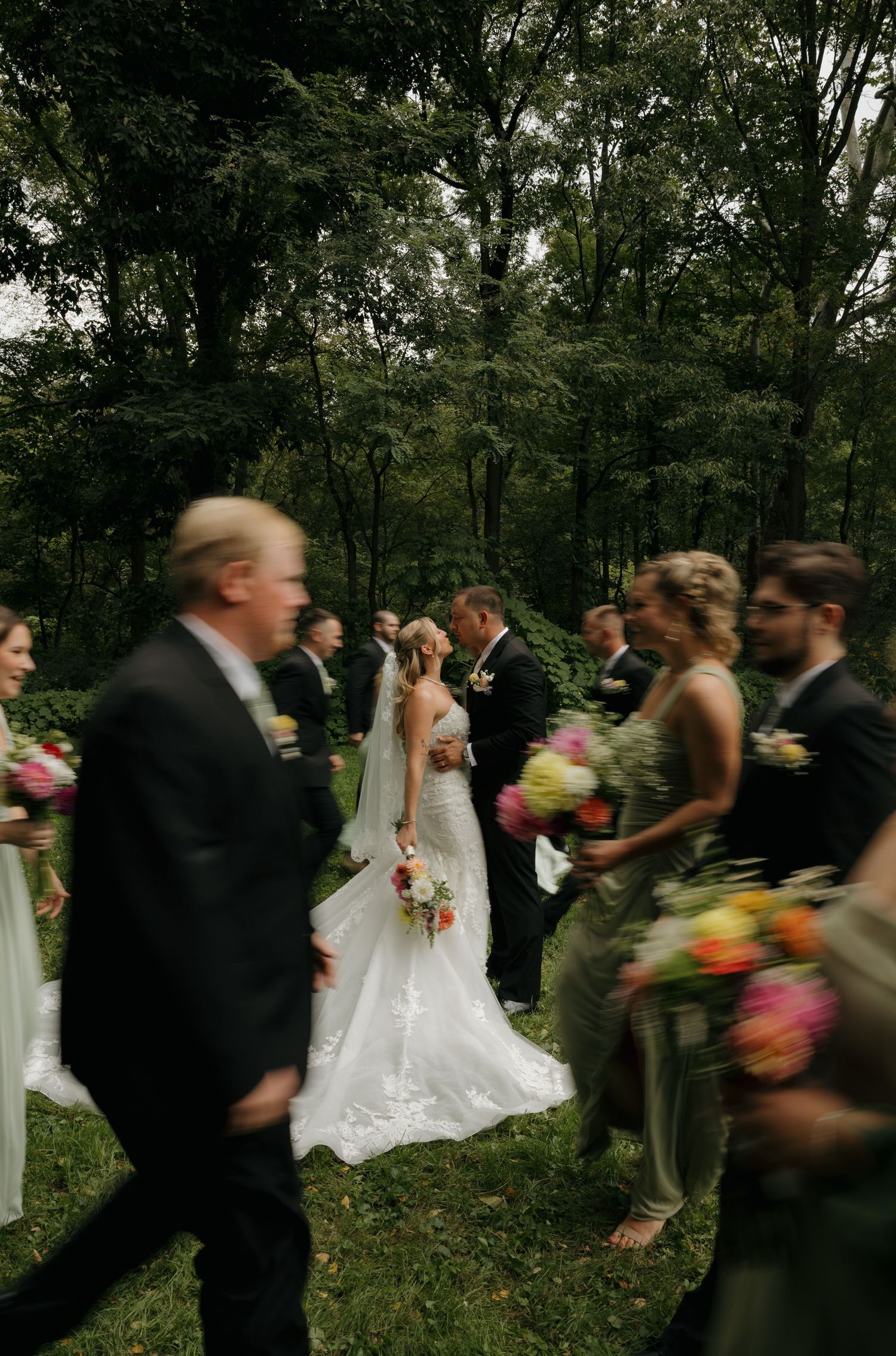 Bride and groom share a kiss surrounded by their wedding party outdoors in a lush green forest setting.