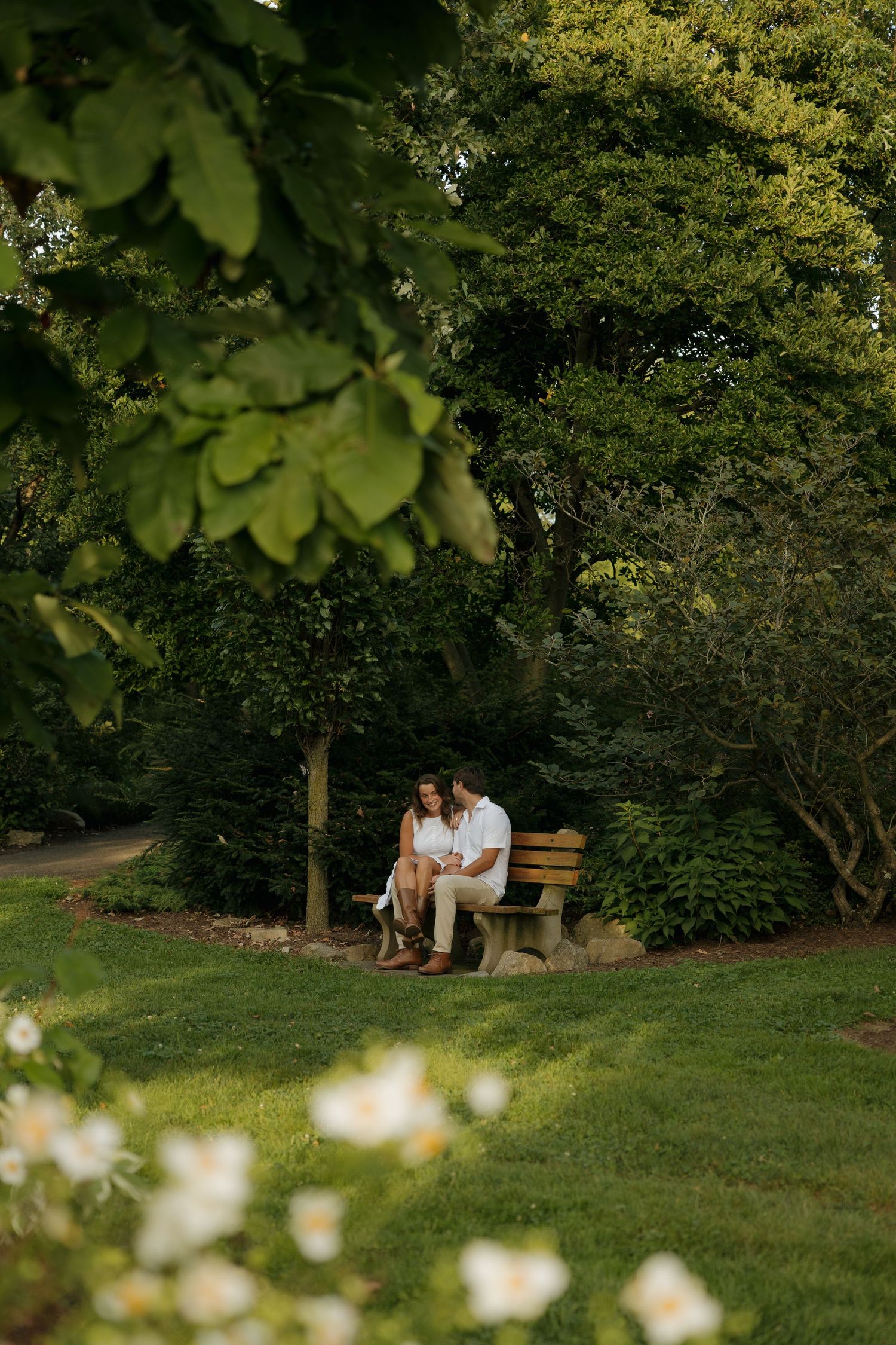 Couple sitting closely on a wooden bench surrounded by lush green trees and white flowers in a serene garden setting.
