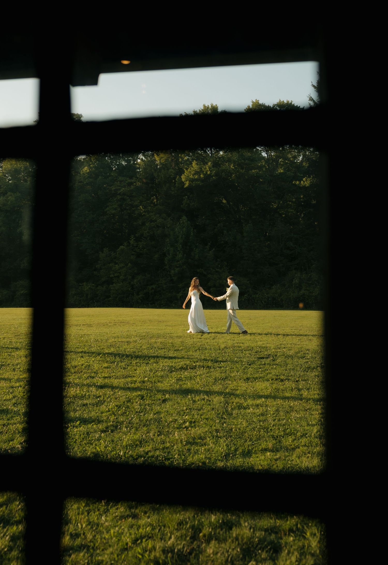 A couple in wedding attire holds hands dancing in a sunlit green field, viewed through a dark window frame.