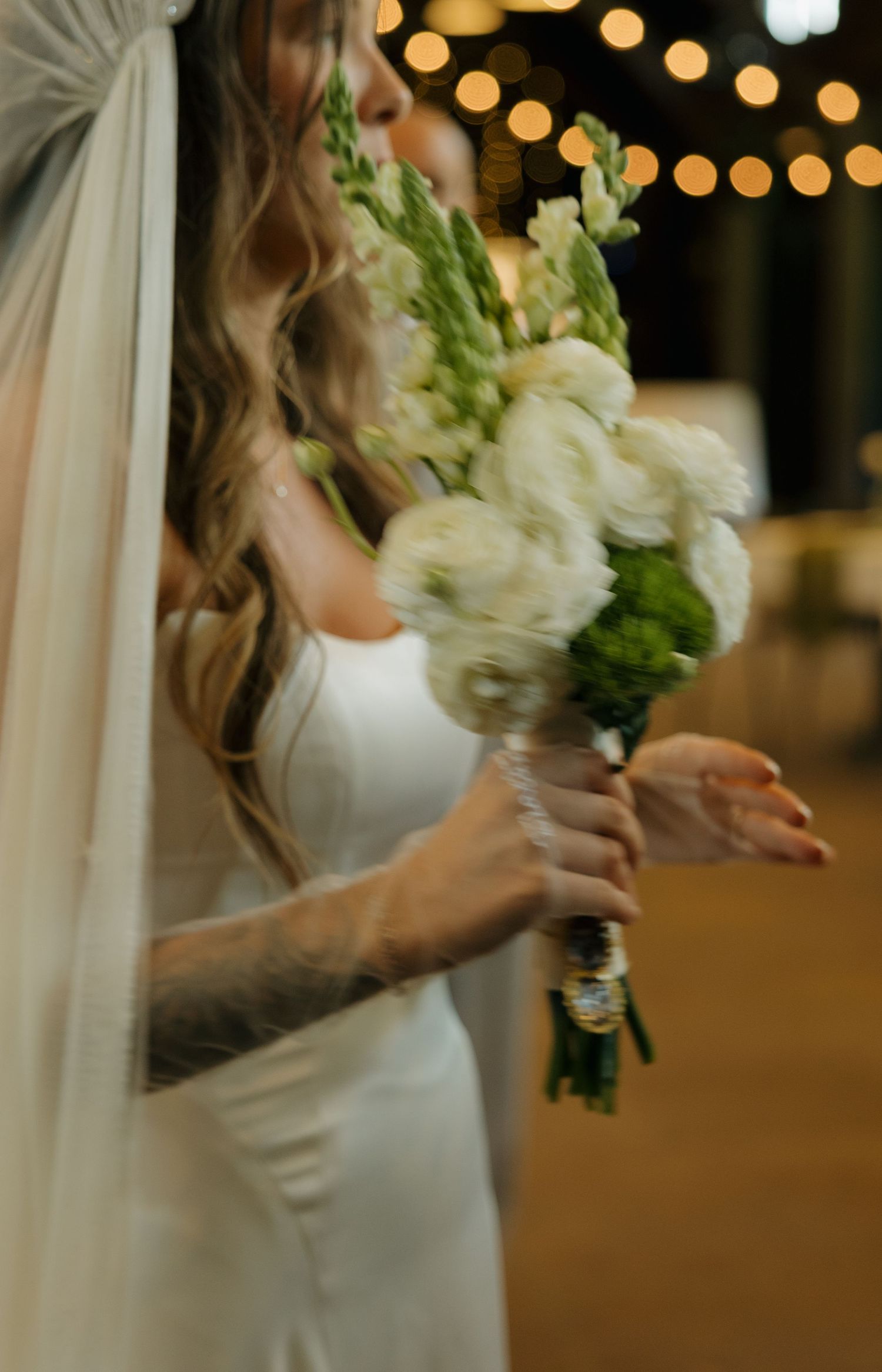 Bride in white gown holding elegant bouquet of white flowers and greenery with bokeh lights in background.