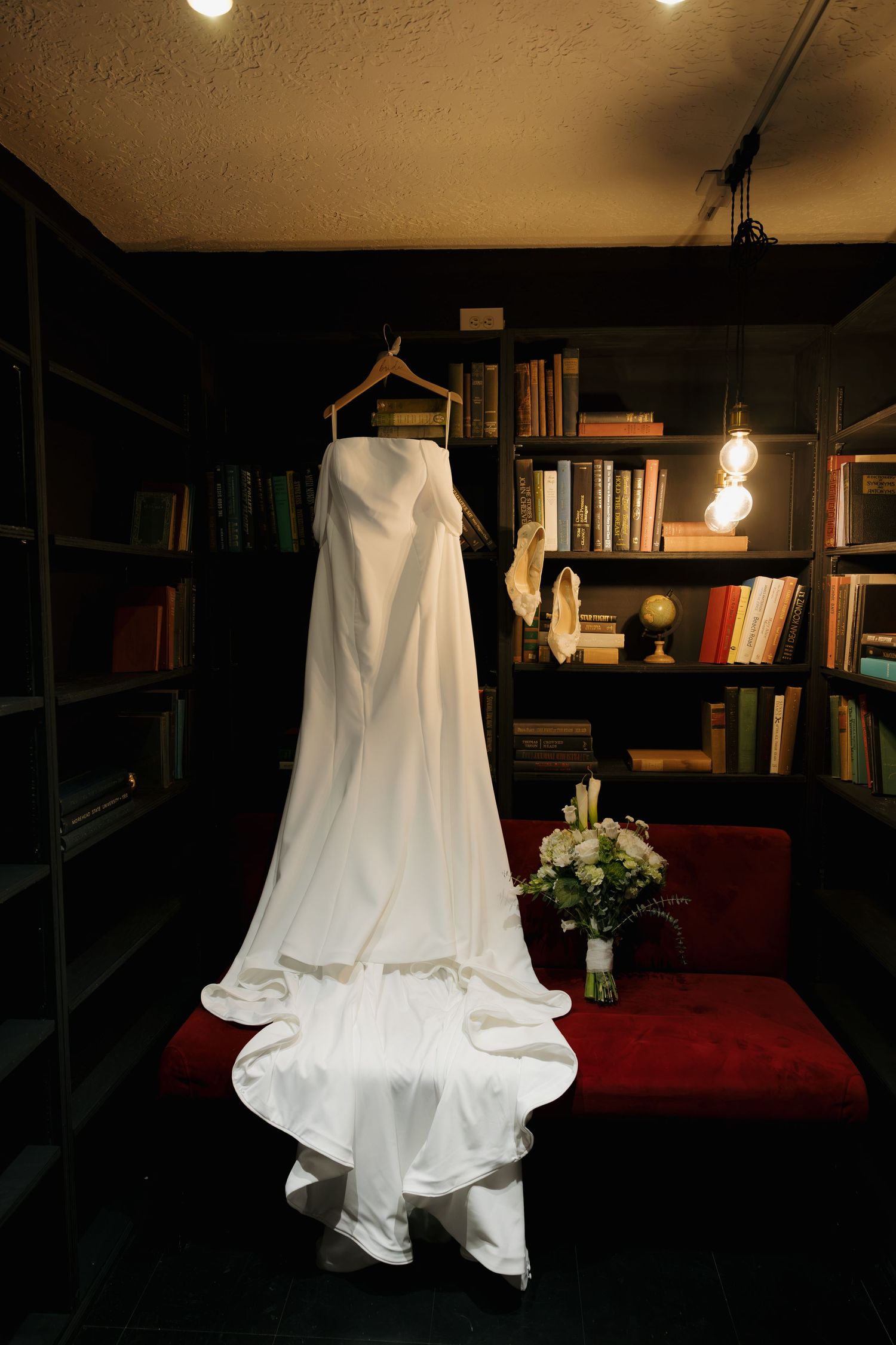 White wedding dress hanging on wooden hanger in cozy library room with bookshelves and bridal bouquet on red chair.