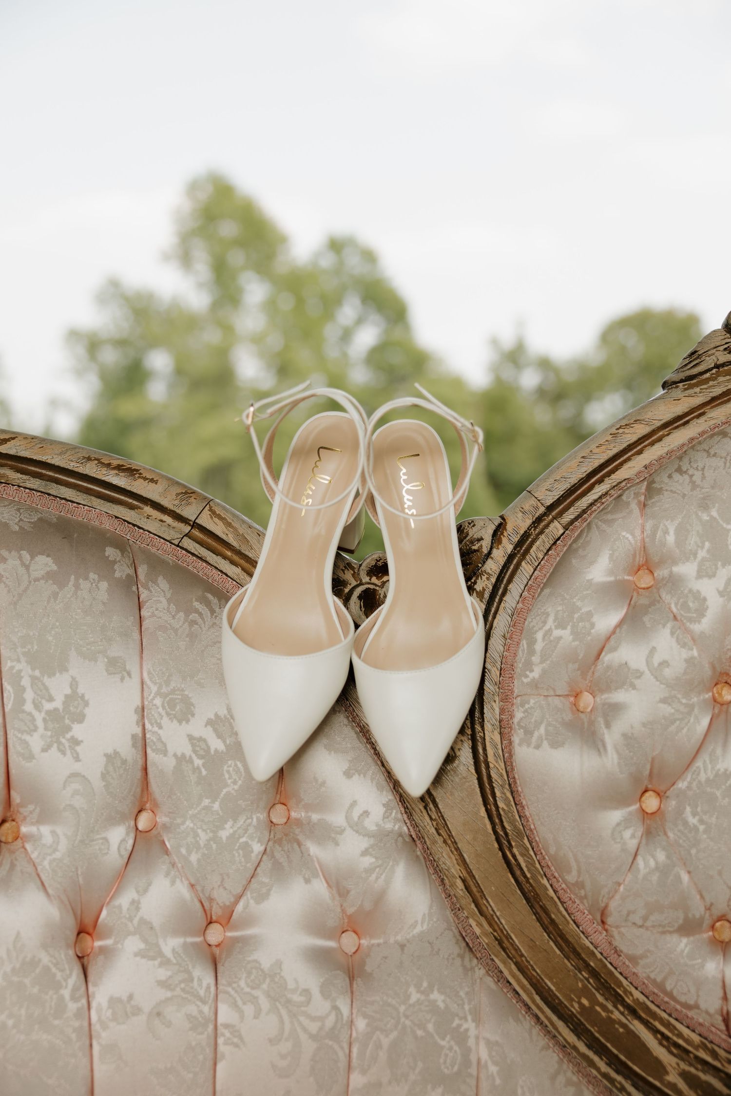 White slingback bridal heels displayed on a vintage tufted ivory settee outdoors with lush greenery in the background.