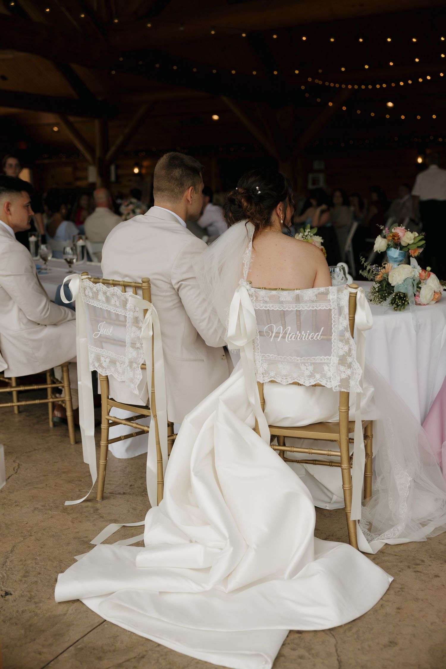 Bride and groom seated at wedding reception sweetheart table with gold chiavari chairs and string lights in barn venue.