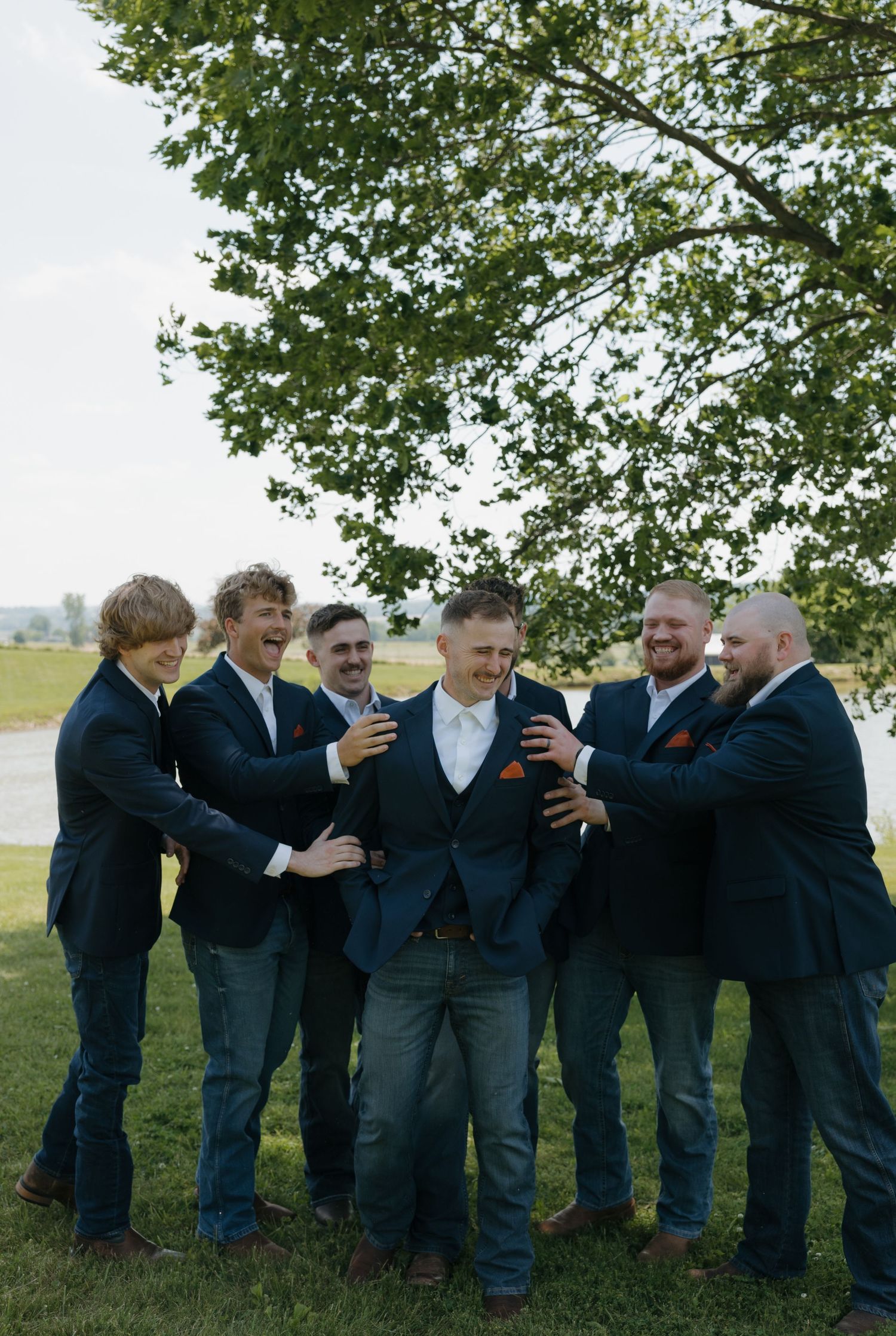 Groom surrounded by laughing groomsmen in navy suits and jeans outdoors under a large green tree.