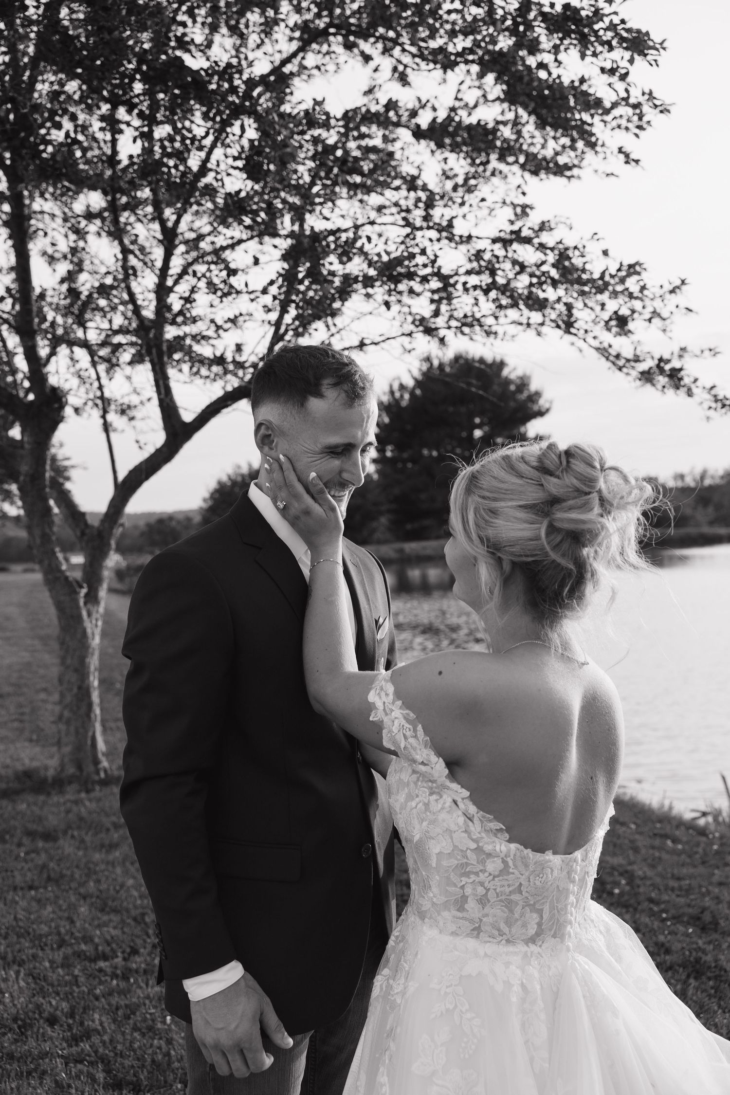 Black and white photo of bride in lace gown tenderly touching grooms face near trees at sunset.