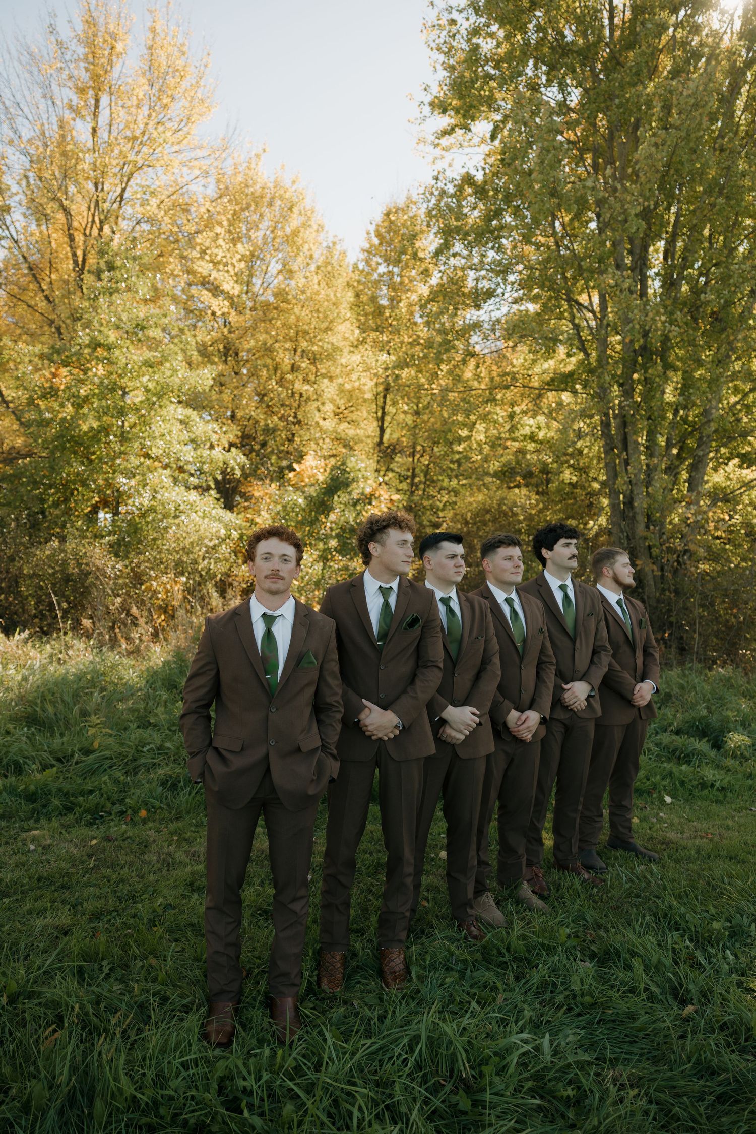 Six groomsmen in brown suits and green ties stand in a line on a grassy field with golden autumn trees behind them.