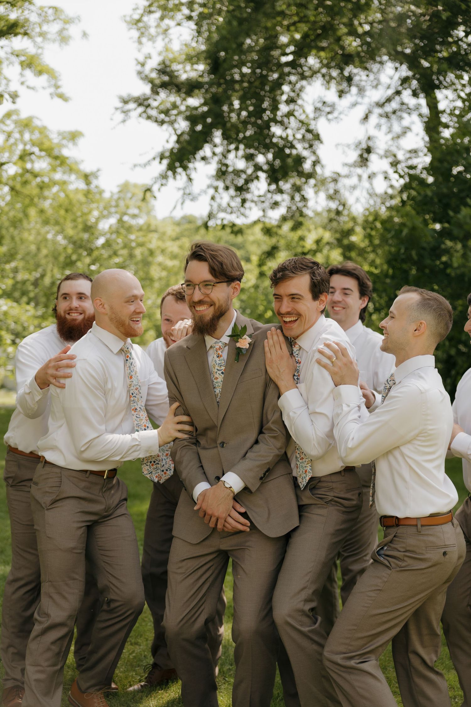 Groom in brown suit laughs with groomsmen in white shirts and floral ties outdoors at a wedding celebration.
