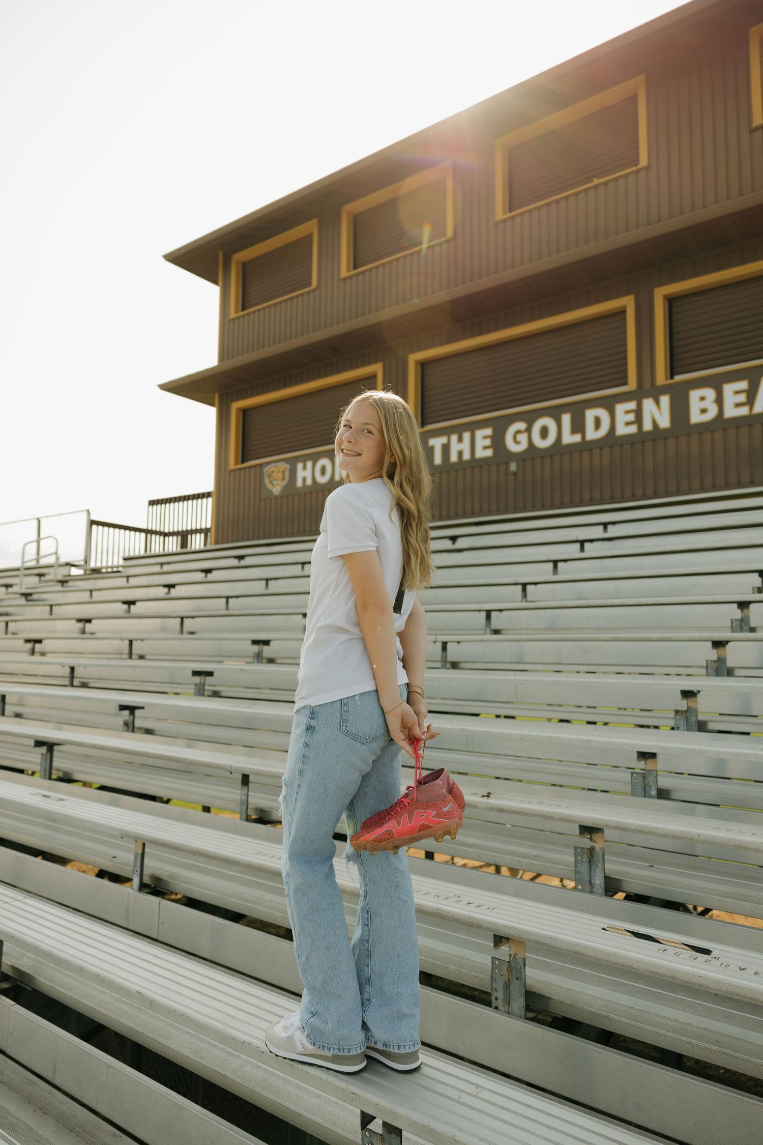 Smiling teenage girl holding red cleats on bleachers at Golden Bears stadium in warm sunlight.