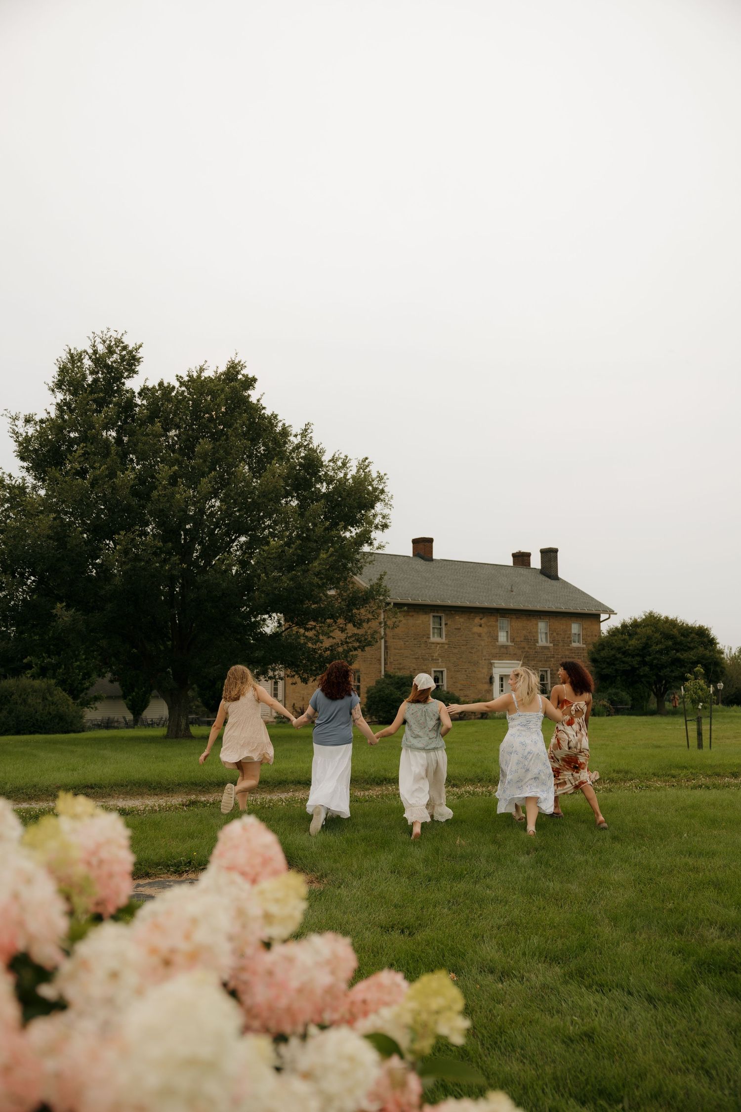 Five women holding hands run across a green lawn toward a brick farmhouse, with pink hydrangeas in the foreground.