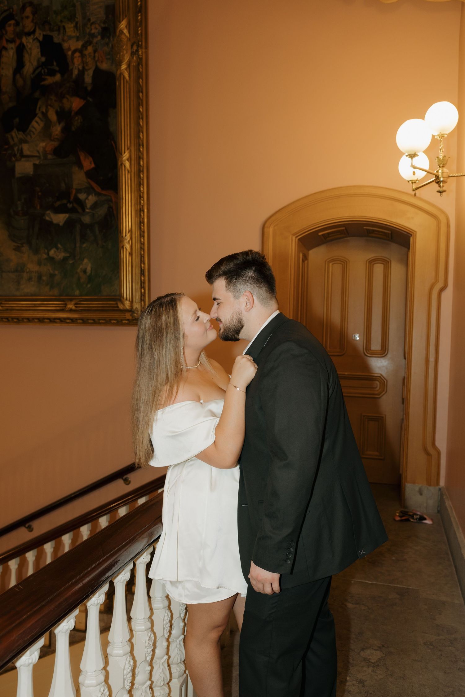 Couple in formal attire sharing a romantic moment on an elegant staircase beside a large painting and wall sconce.