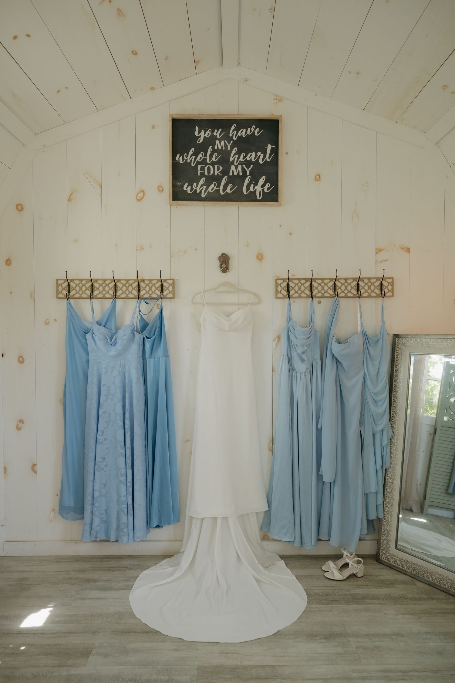 White wedding gown and blue bridesmaid dresses hanging on rustic wall hooks beneath a romantic chalkboard sign in a bridal suite.